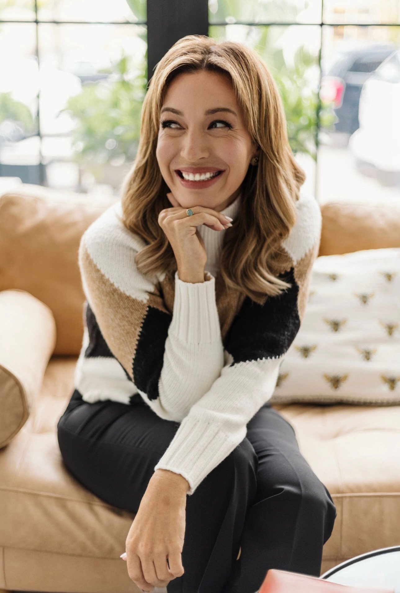 A woman with wavy, shoulder-length blonde hair smiling and looking to her left, sitting on a tan leather sofa in a well-lit room with large windows.