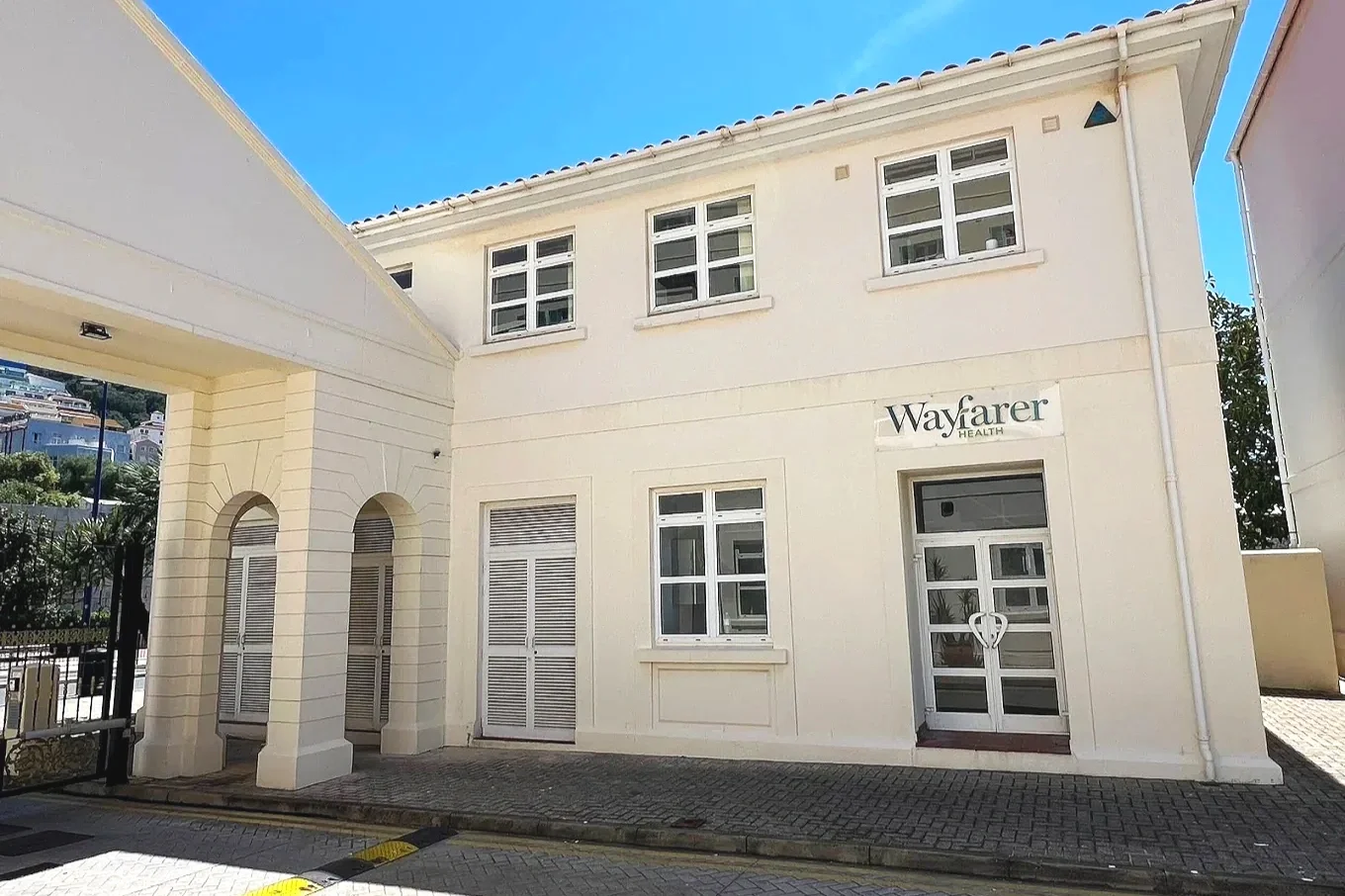 Exterior view of a white two-story building with a sign that reads 'Wayfarer Health.' The building has multiple windows, and the front entrance features glass double doors with a heart-shaped handle. There is a paved sidewalk and a curb in front of the building.