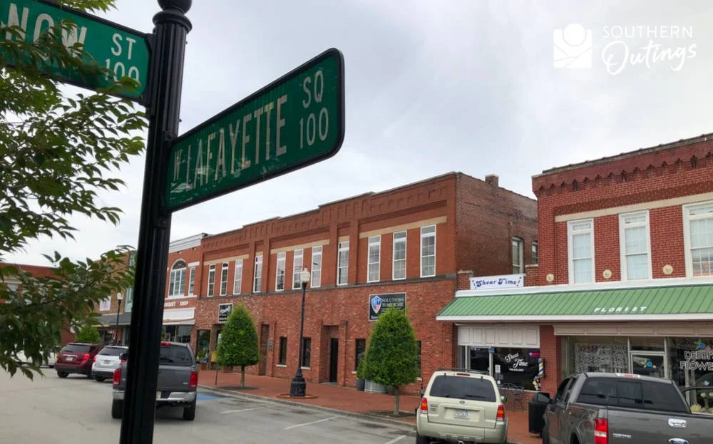 Street signs for Lafayette Square and W. H. St, with a view of brick buildings and parked cars along the street.
