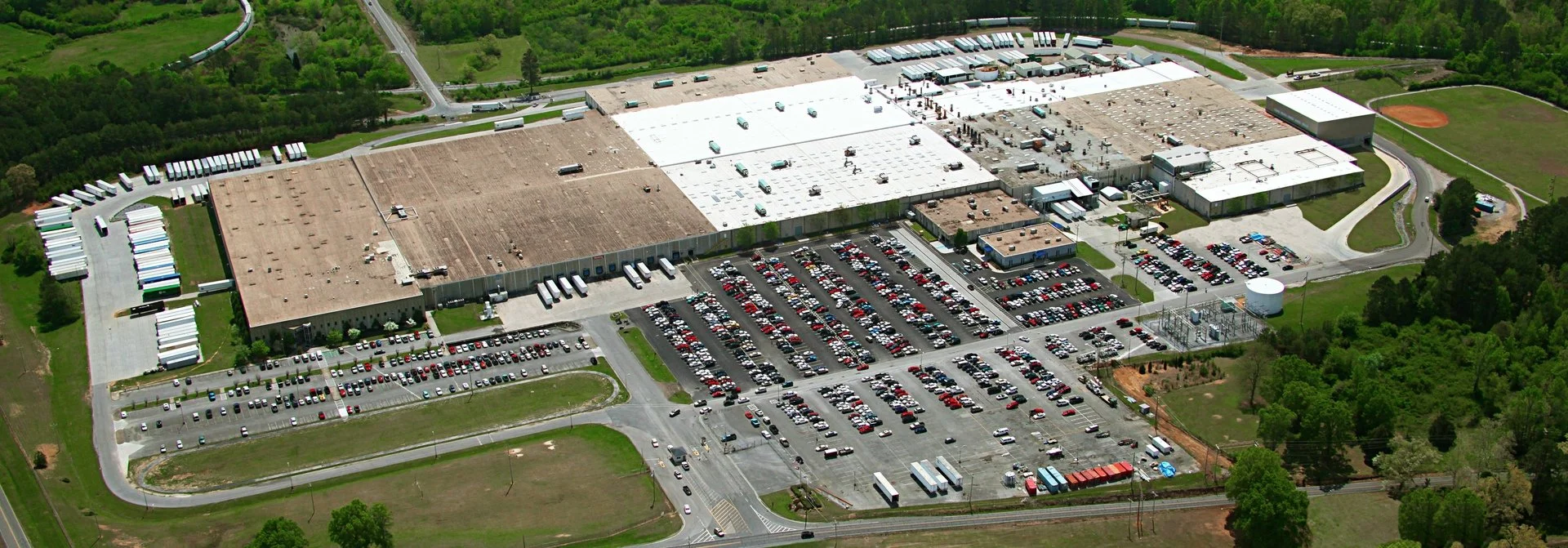 A large warehouse or factory building with numerous parked cars and trucks surrounding it, set in a green landscape with trees and fields.