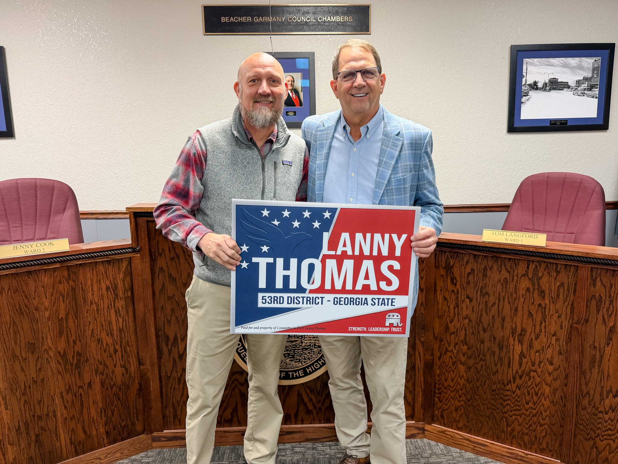 Two men smiling at a political event, holding a campaign sign for Lanny Thomas for Georgia State, 53rd District, in a council chambers room. The man on the left wears a plaid shirt with a gray vest, and the man on the right wears a blue plaid blazer. There are nameplates on the desks behind them saying 'Jenny Cook, Ward 2' and 'Tom Langford, Ward 1', with framed pictures on the wall.