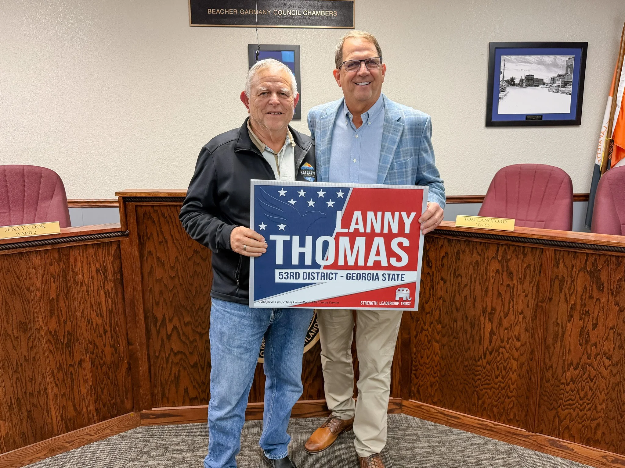 Two men standing in a government chamber, holding a campaign sign for Lanny Thomas. The man on the left wears a black jacket and jeans, the man on the right wears a light blue plaid blazer and beige pants. Behind them are wooden chairs and a wall with framed photos and a nameplate.
