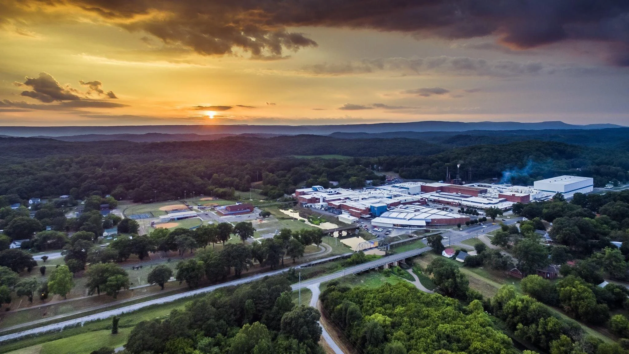 Aerial view of a small town surrounded by lush green trees, with a school and a factory in the distance at sunset.