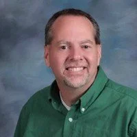 Man with short hair, wearing a green button-up shirt, smiling against a blue-gray background.