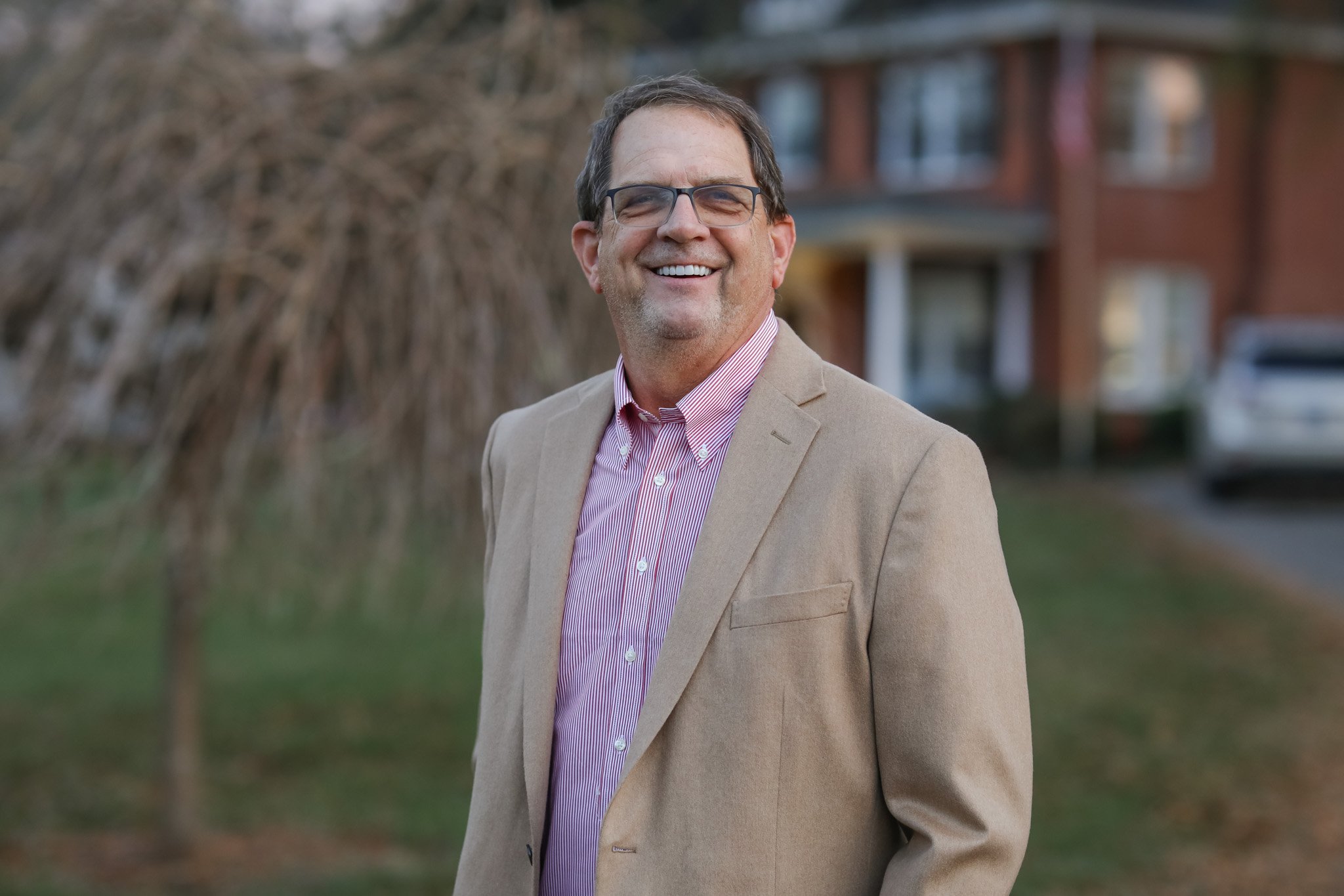 A smiling man with glasses, wearing a beige blazer and pink striped shirt, standing outdoors in front of a house with a tree and a parked car in the background.