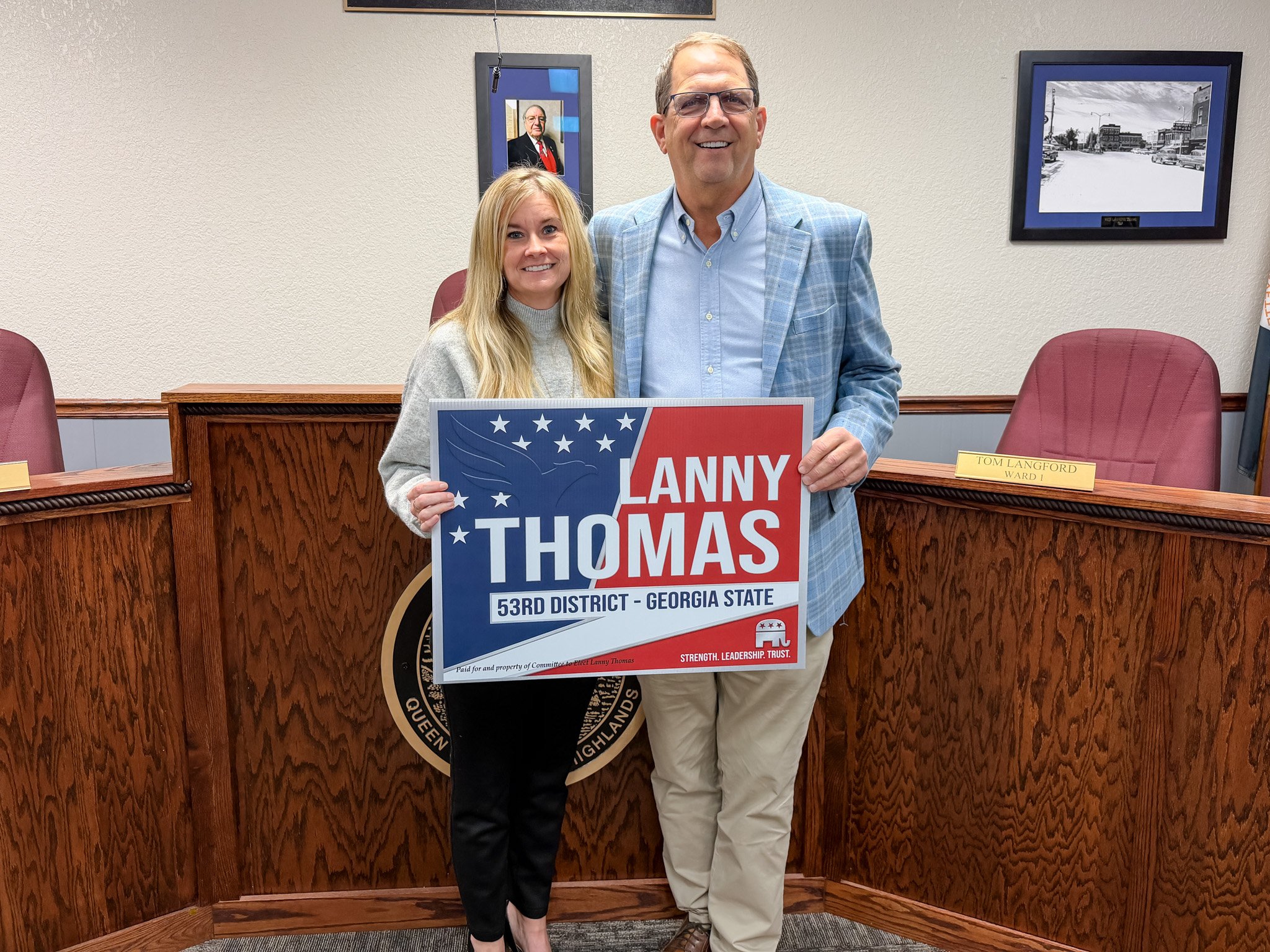A man and a woman standing behind a wooden podium, holding a campaign sign for Lanny Thomas for Georgia State 53rd District. The man wears glasses and a blue plaid jacket, and the woman has long blonde hair and is wearing a gray sweater. The setting appears to be a formal meeting room with framed pictures on the wall.