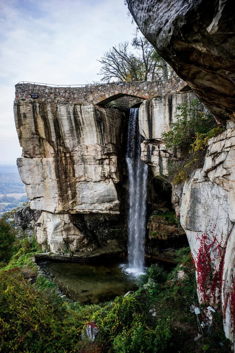 A waterfall flowing from a rock formation with an arched bridge above it, surrounded by trees and greenery in a natural landscape.