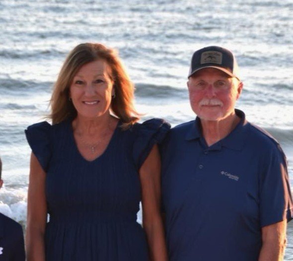 A smiling woman and an older man standing side by side on a beach with ocean waves in the background.