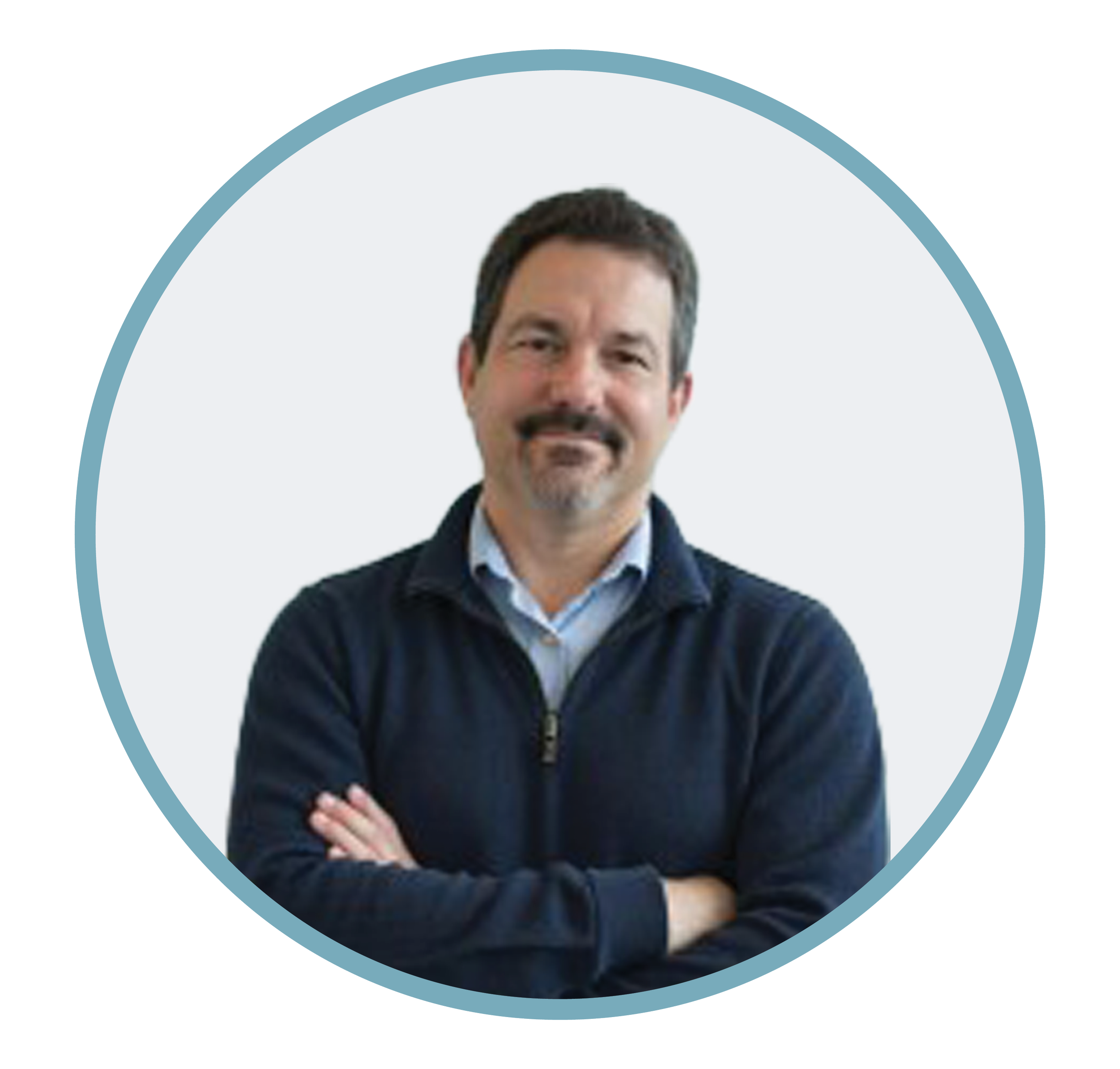 A man with dark hair and a beard, wearing a dark jacket over a light blue shirt, standing with arms crossed against a plain white background.
