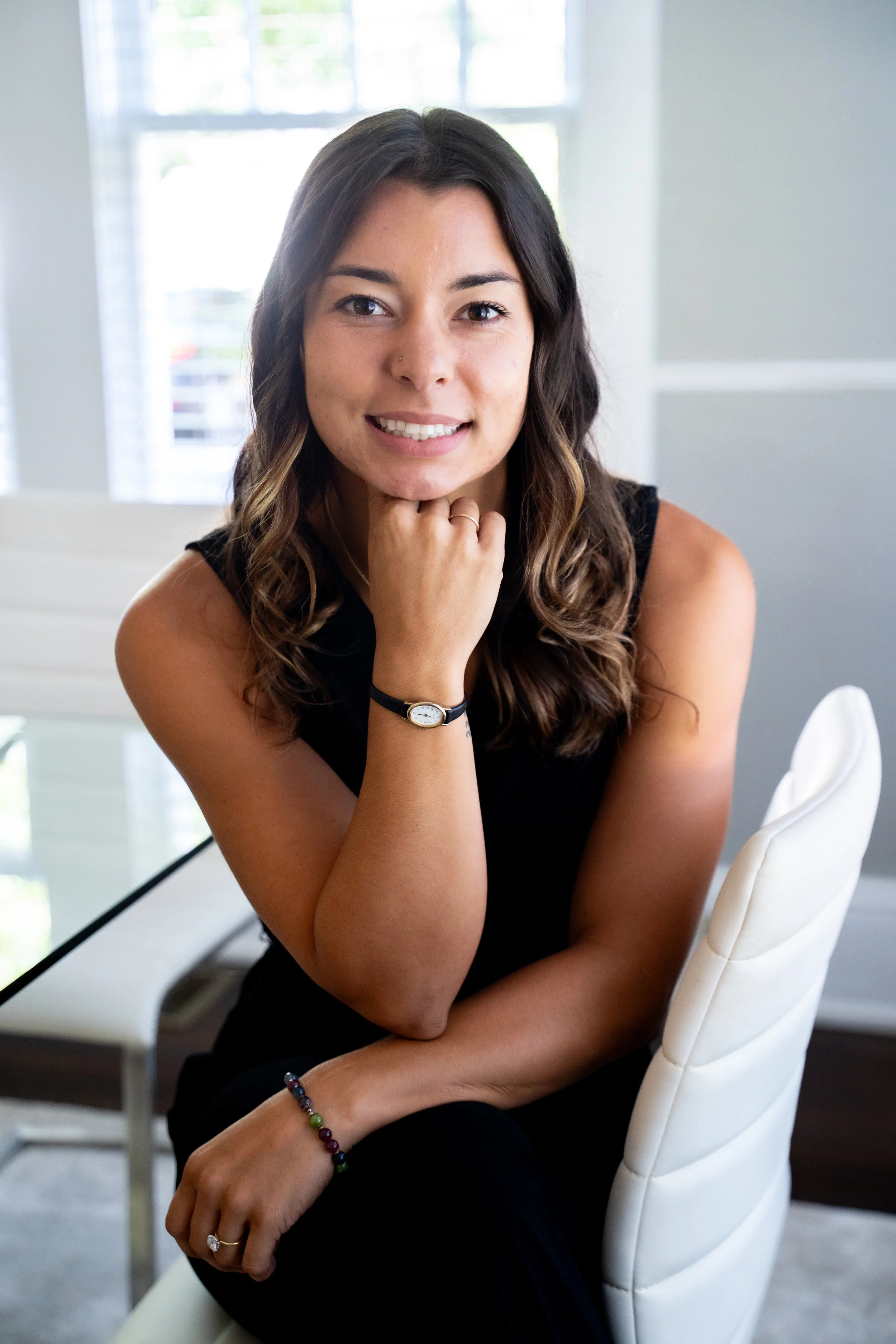 Young woman with long wavy hair, wearing a black sleeveless top, smiling, sitting on a white chair in a bright room with large windows.