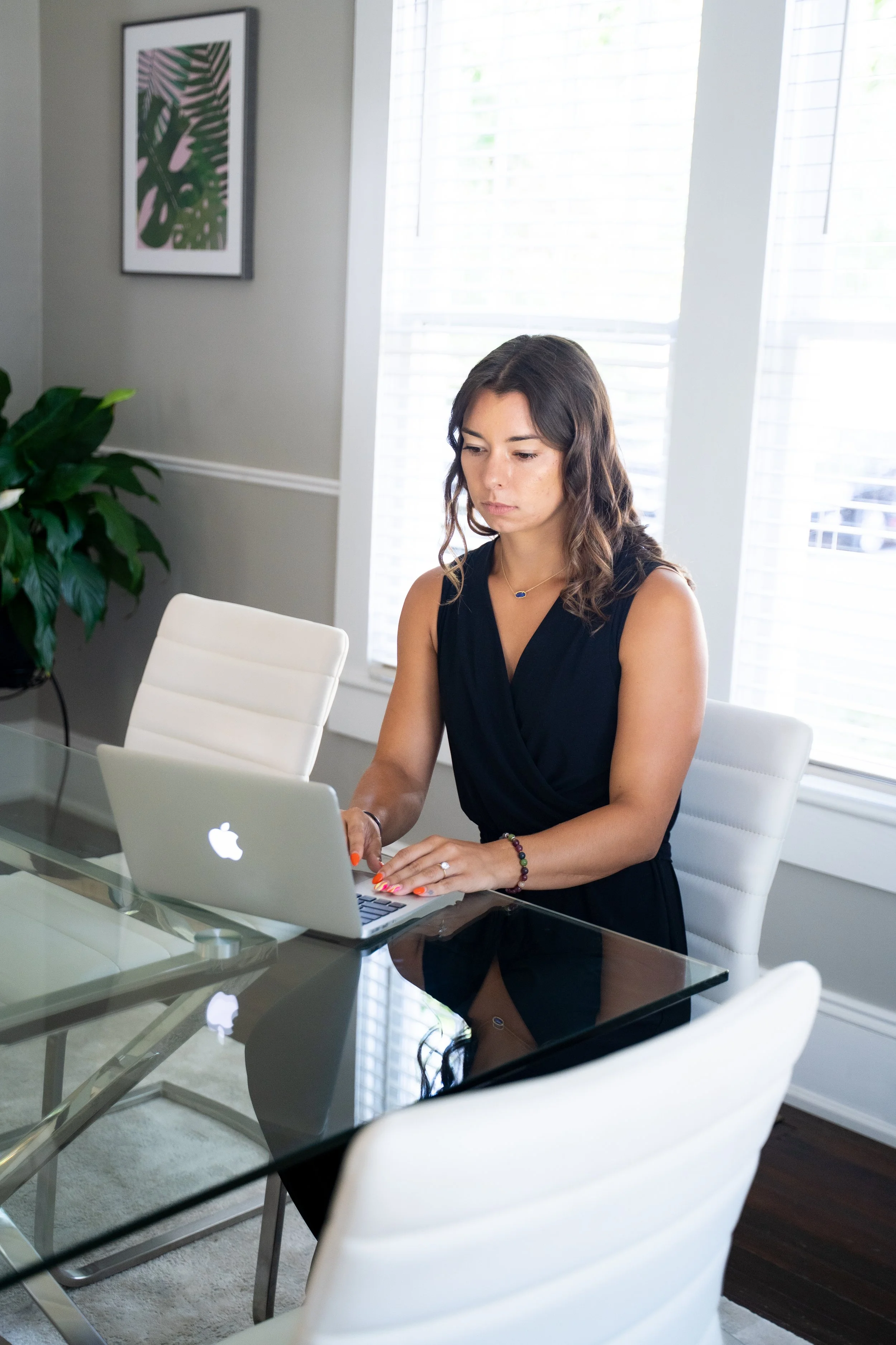 A woman with wavy brown hair working on a silver MacBook laptop at a glass table in a bright, modern office or home space with natural light from large windows.
