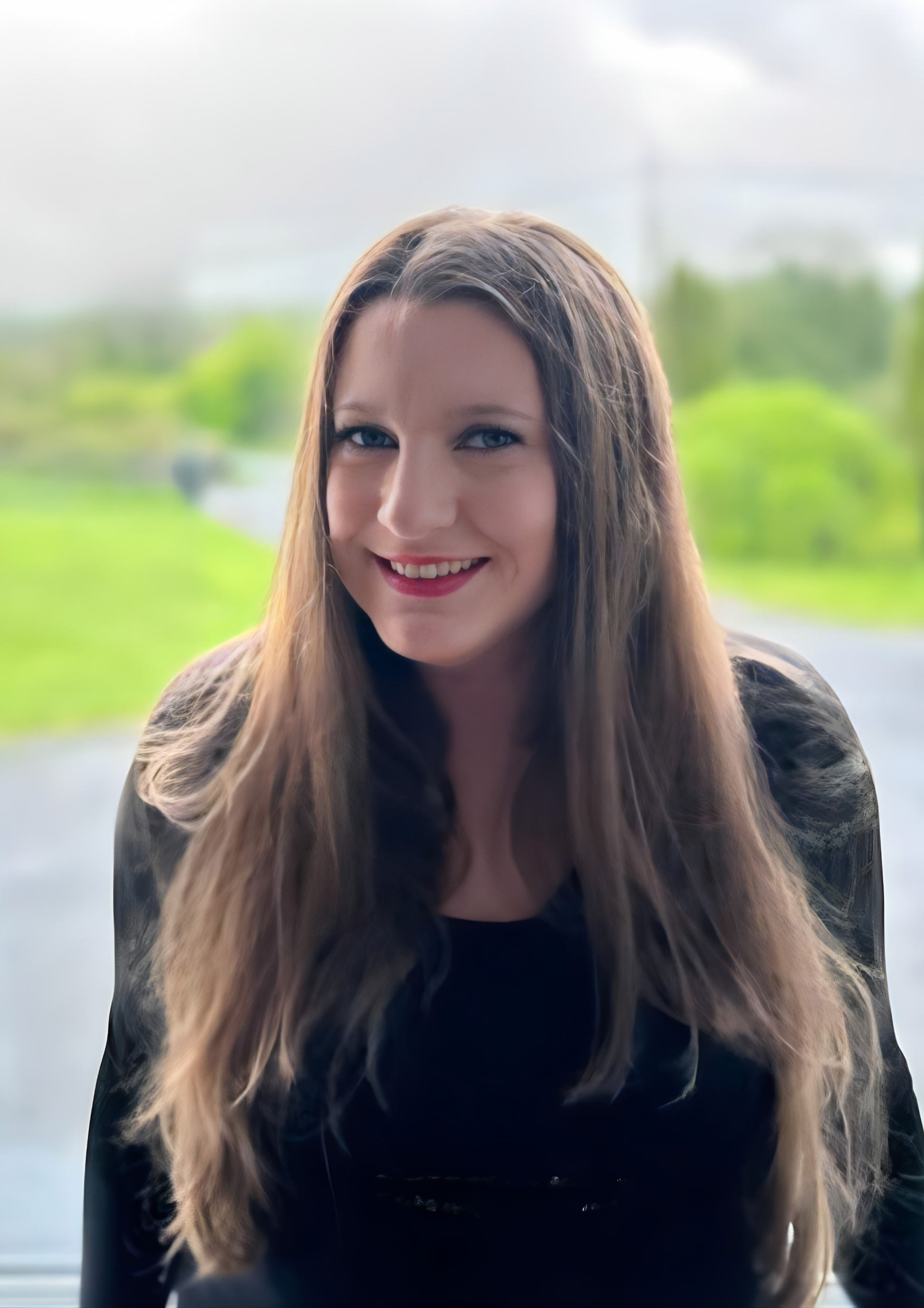Portrait of a woman with long, wavy brown hair, smiling and looking at the camera, with a blurry outdoor background.