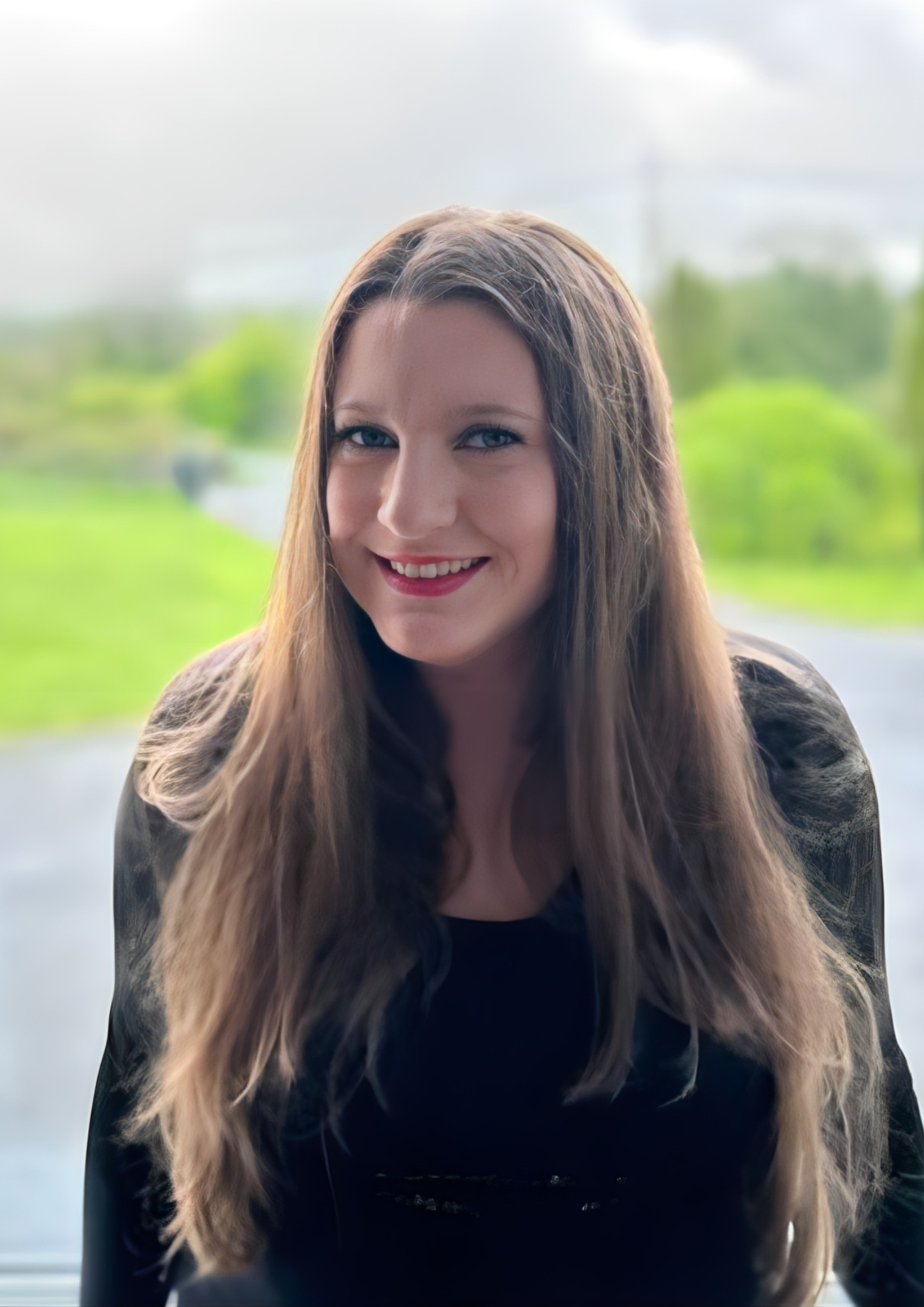 A smiling woman with long, wavy dark blond hair and blue eyes, wearing a black top, standing in front of a window with a blurred green outdoor landscape in the background. She is also a certified nutritionist and gluten free diet expert.