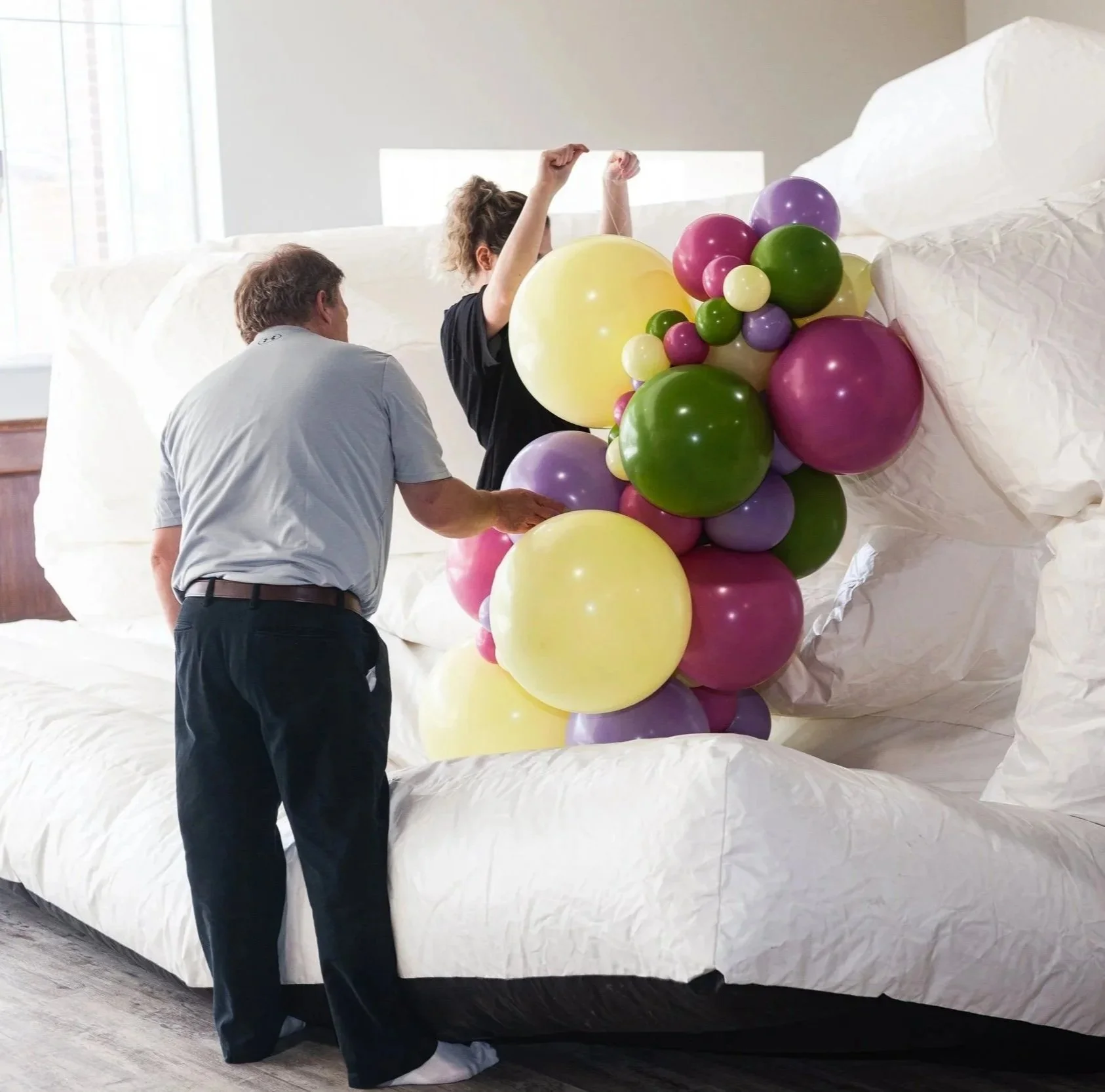 People inflate a large cluster of colorful balloons inside a room with a white couch.