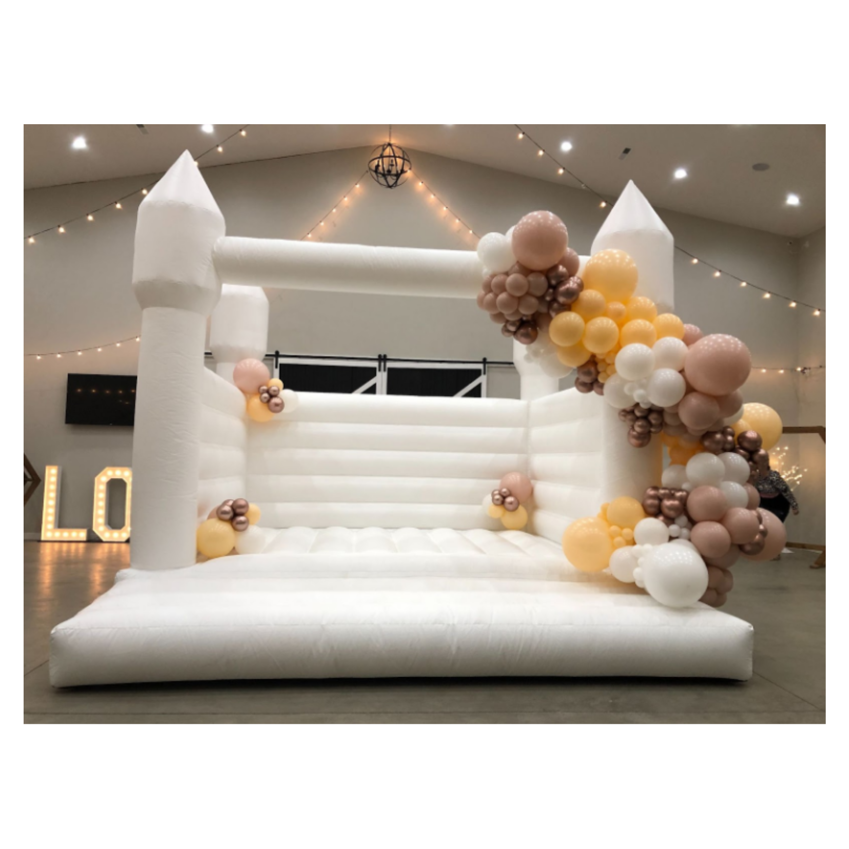 A white inflatable castle decorated with pastel balloons in beige, peach, white, champagne, and brown, arranged in clusters at the corners and sides, set up indoors with string lights and a lit letter sign in the background.