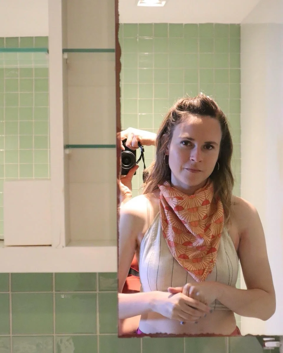 Sammie Downing taking a mirror selfie in a bathroom with green tile walls, wearing a striped top and red bandana, with a person partially visible behind her.