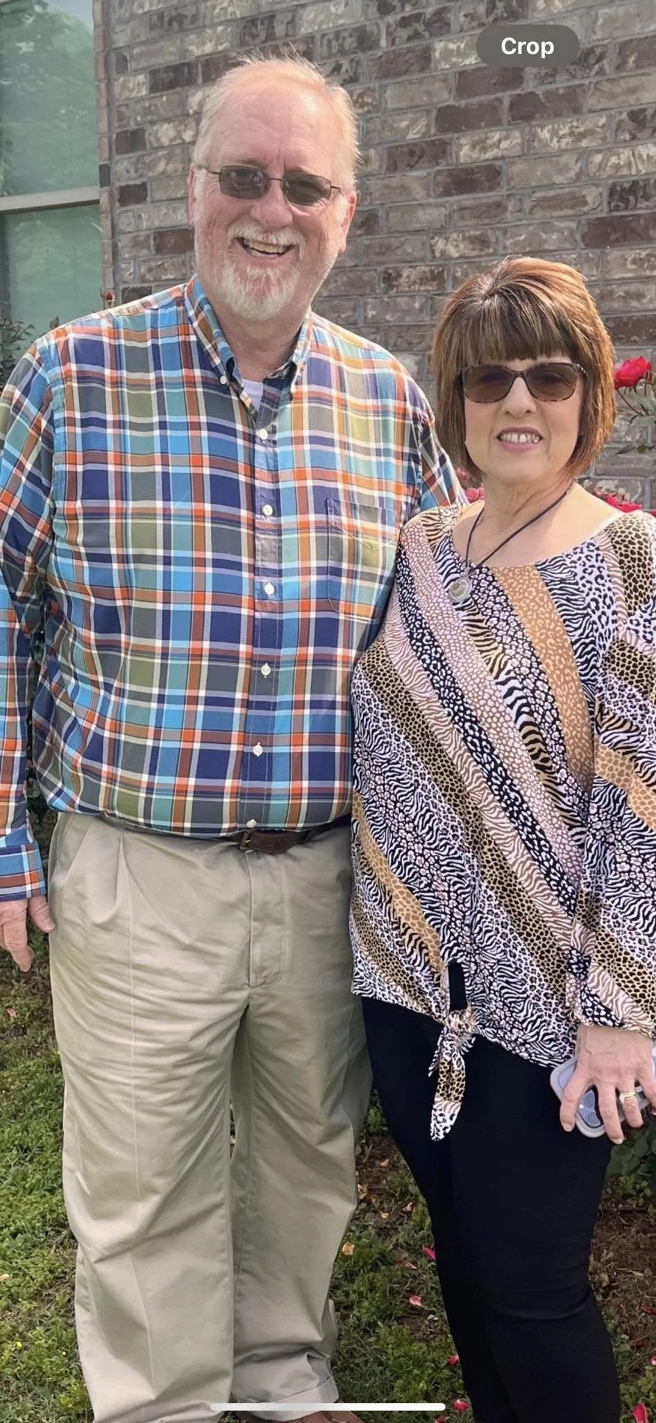 A smiling man and woman standing outdoors in front of a brick wall, with the man wearing a colorful plaid shirt and khaki pants, and the woman wearing a patterned blouse, black pants, and sunglasses.