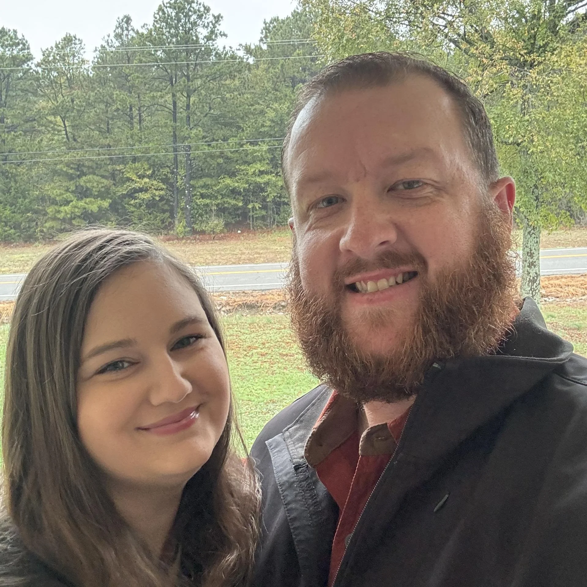 A man with a beard and a young girl are smiling for a selfie outdoors with trees and a road in the background.