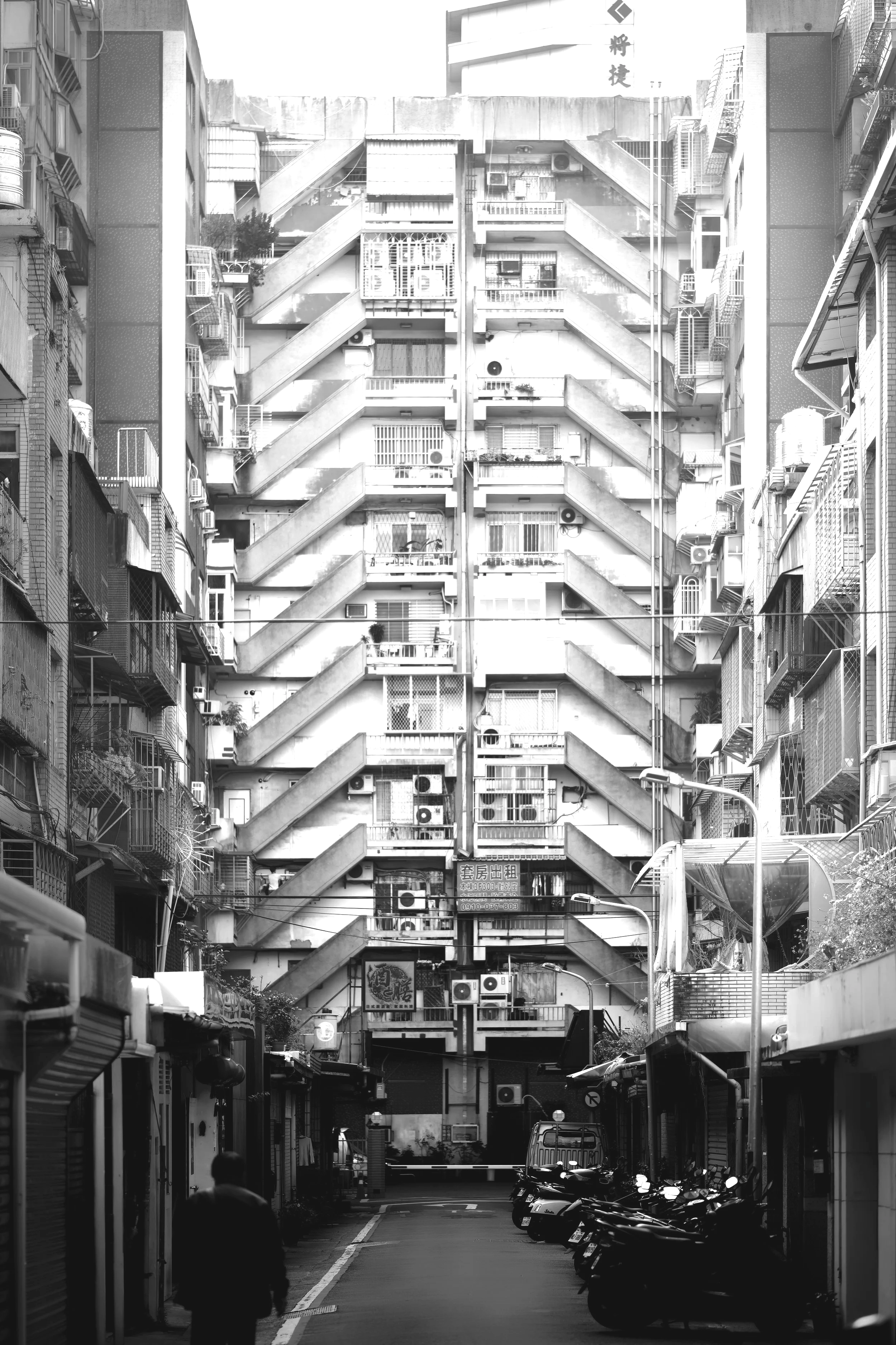 Black and white photo of a narrow alleyway flanked by tall apartment buildings with external staircases. Motorcycles are parked along the street, and a person is walking away from the camera. Air conditioning units and balconies are visible on the buildings.