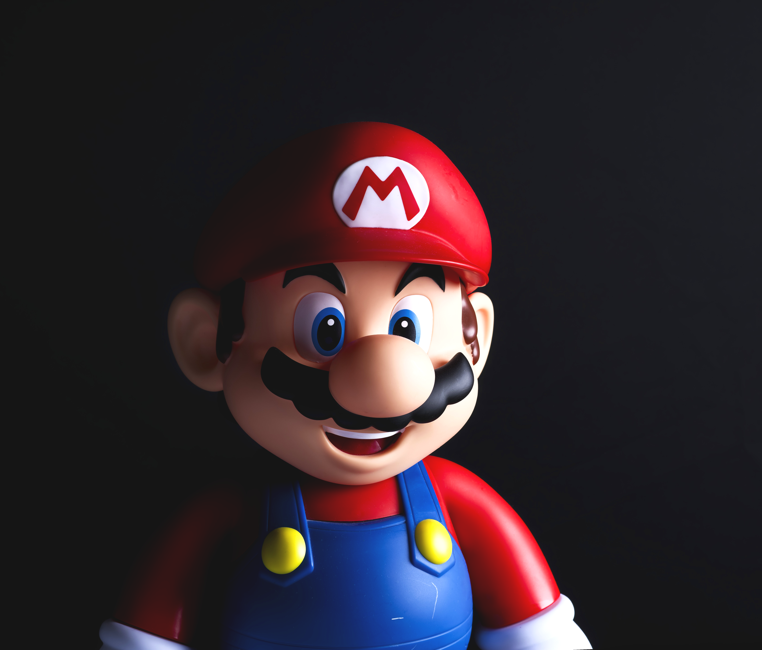 A close-up of a Mario figurine with a red hat, blue overalls, and a big mustache, set against a dark background.