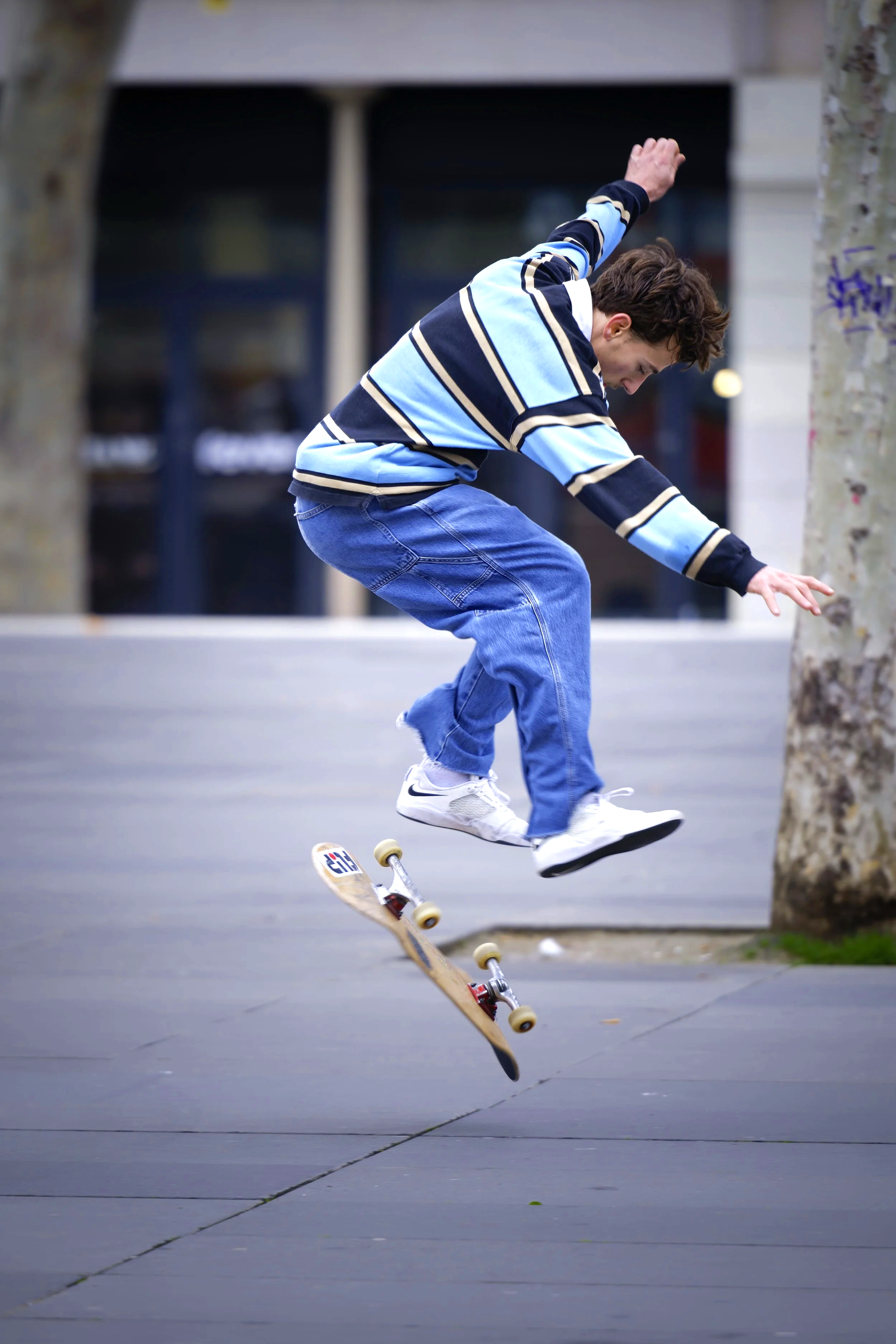 A young man wearing a striped sweater and jeans skateboarding outdoors next to a tree.