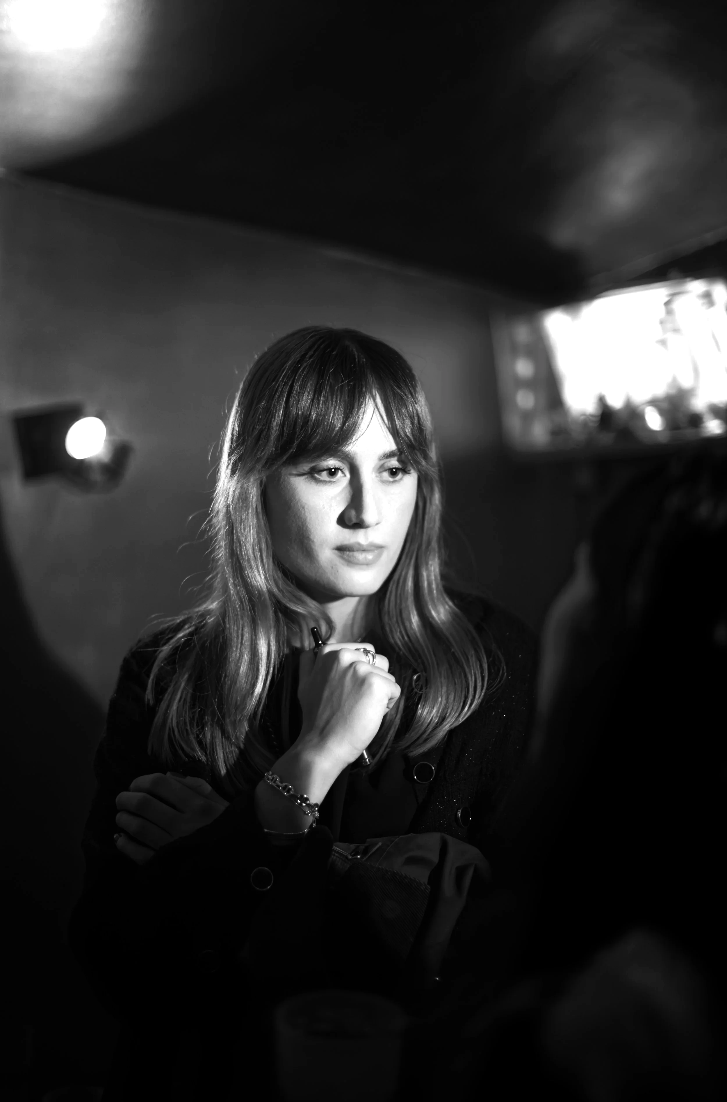 A black and white photo of a woman with long hair, looking off to the side with a contemplative expression, sitting in a dimly lit room.