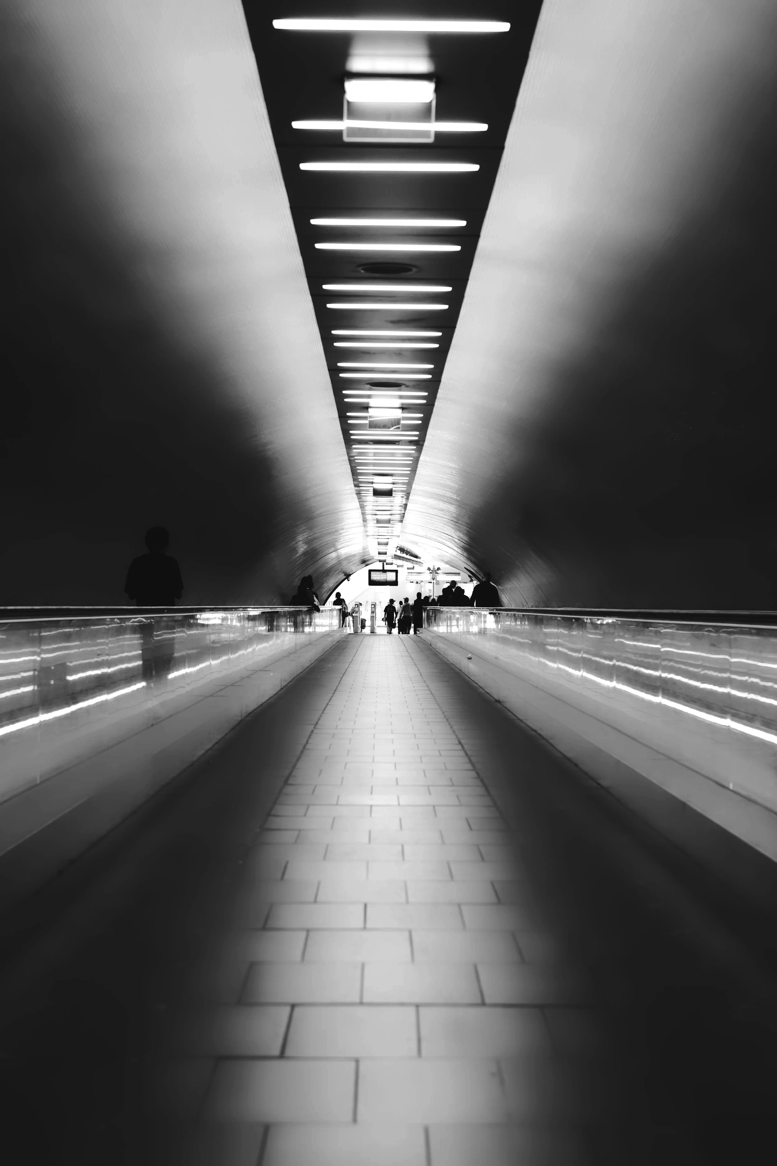A black-and-white photo of an underground walkway with a ceiling lined with evenly spaced fluorescent lights, leading to an exit with silhouettes of people walking.