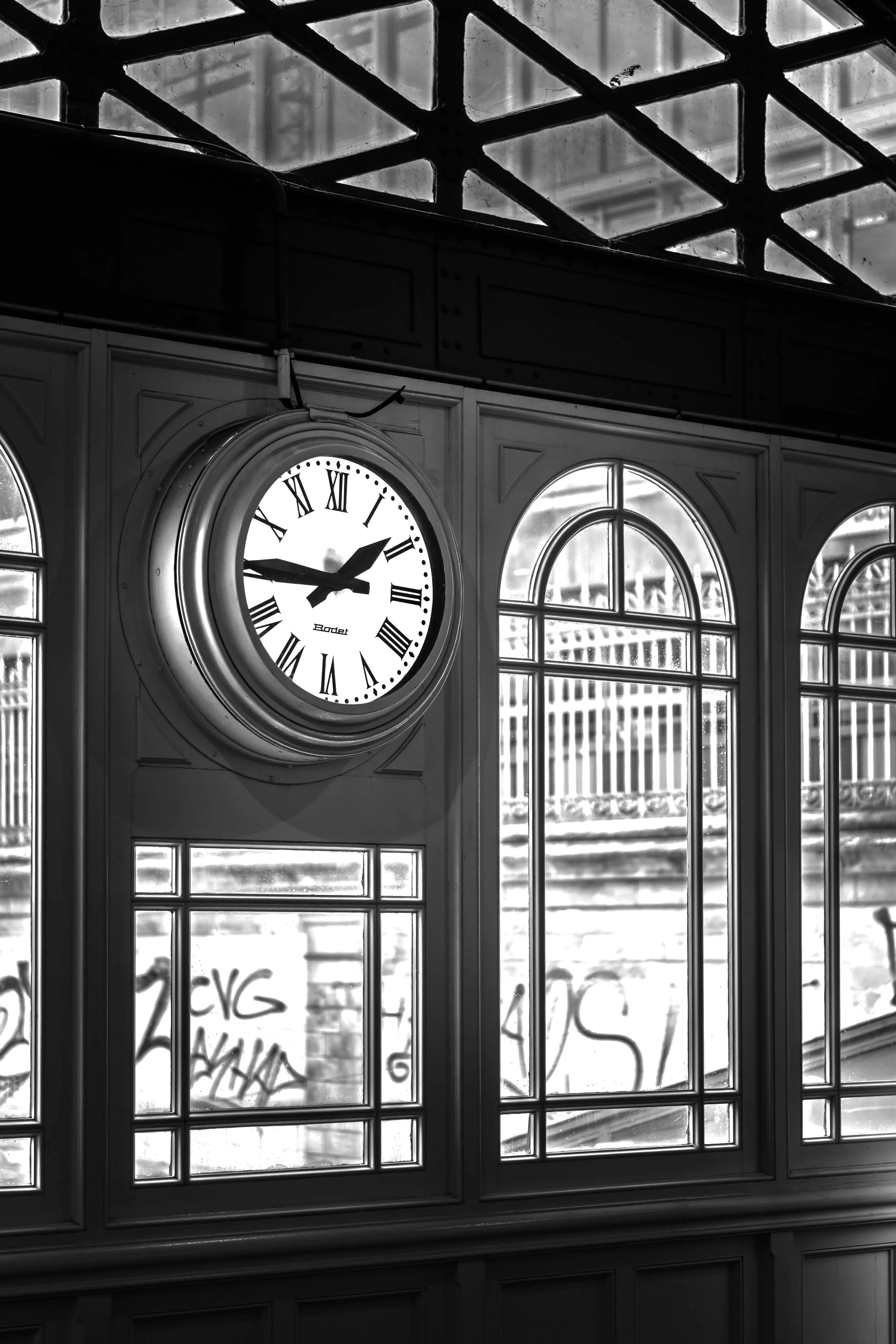 A black and white photo of a train station interior with a round clock showing 2:13, above decorative windows with graffiti outside.