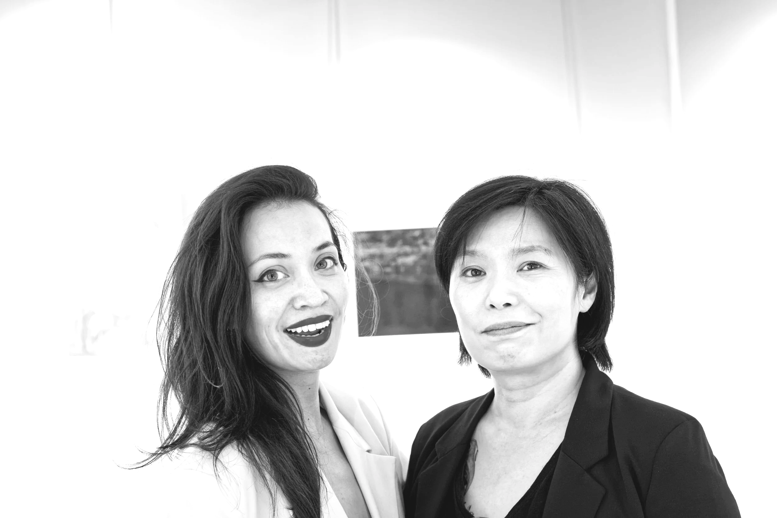 Two women posing together in a photo, one with long dark hair and the other with short dark hair, both dressed in professional attire, indoors with a bright background and a small framed picture on the wall.