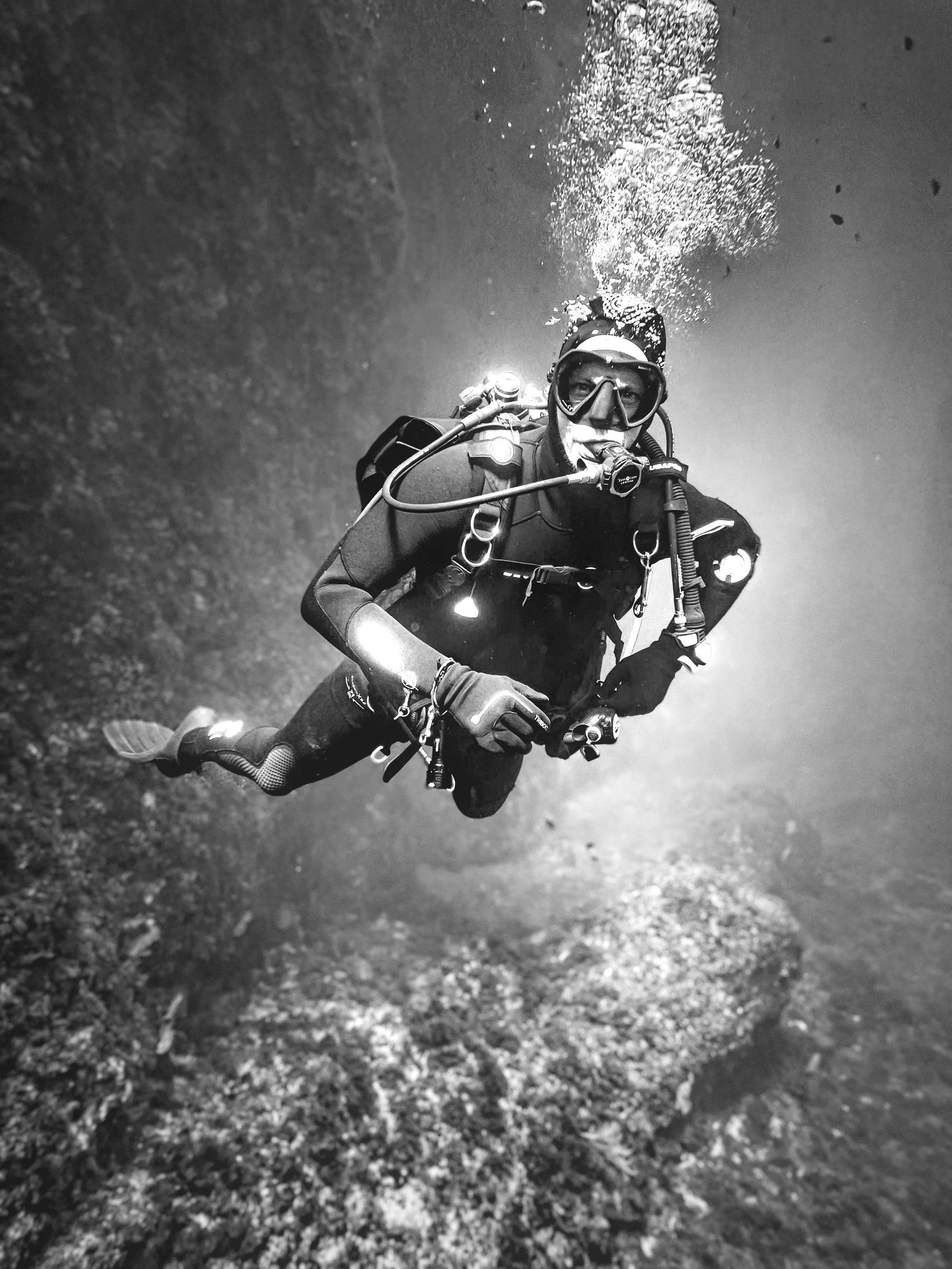Black and white photo of a diver underwater wearing scuba gear, including mask, snorkel, wetsuit, and fins, holding a camera, with bubbles rising above.