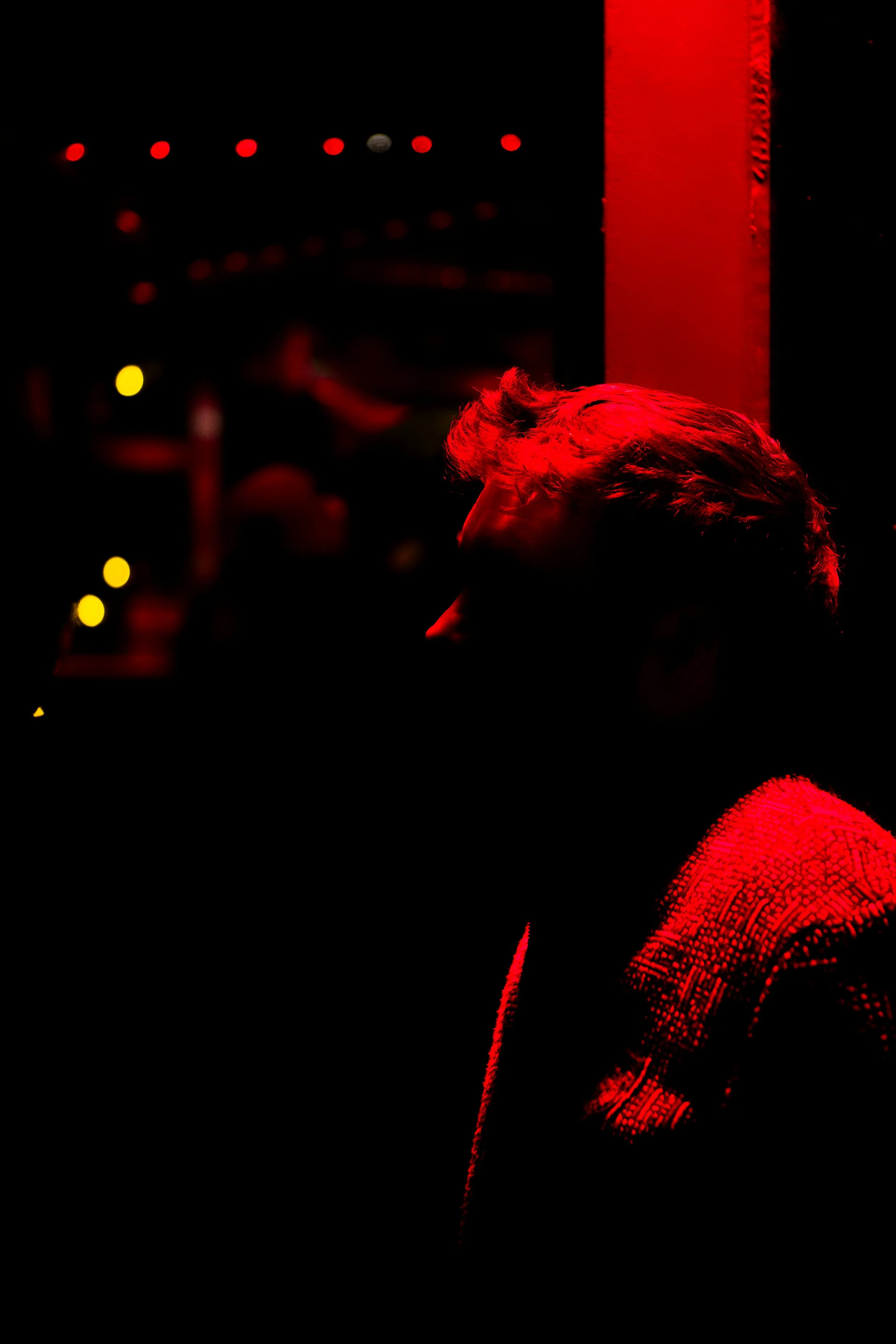 A person with light-colored hair illuminated by red lighting, standing against a dark background with blurred yellow and red lights.
