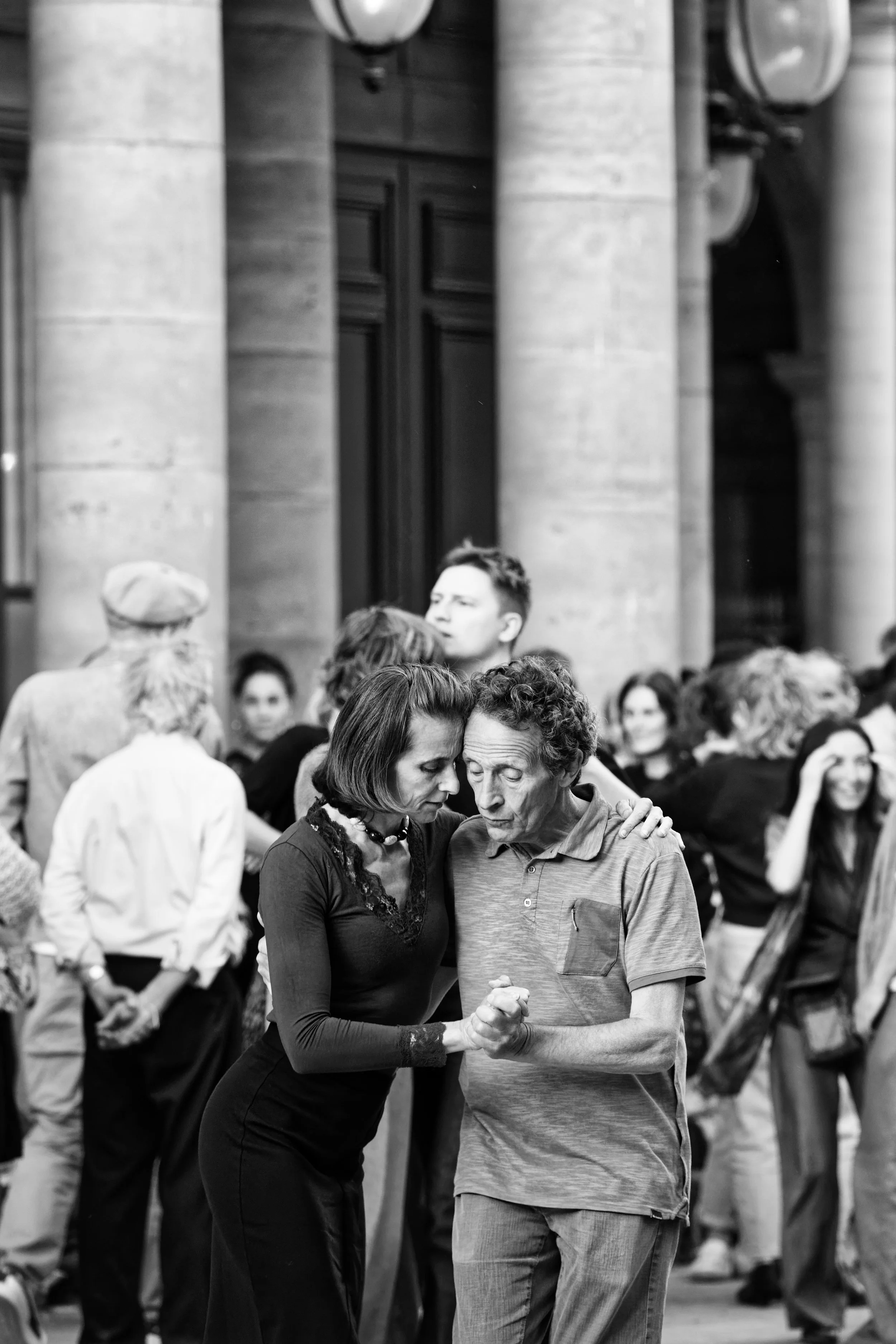 Black and white photo of a woman and man dancing closely together with their eyes closed, surrounded by people in an outdoor setting with large columns in the background.