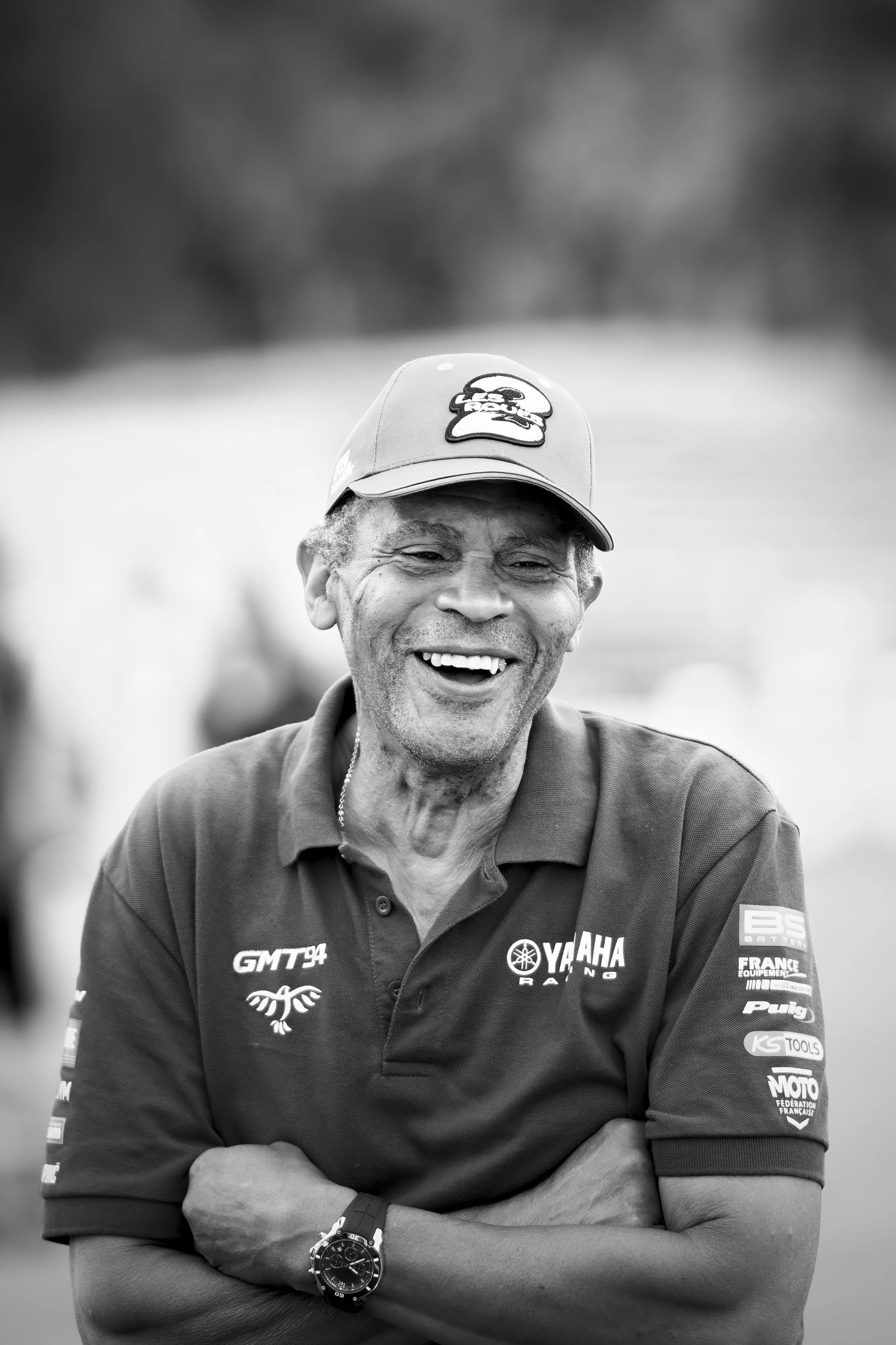 A black and white photo of a smiling man wearing a yamaha racing polo shirt, a cap with a cartoon face, and a watch on his wrist, with arms crossed.
