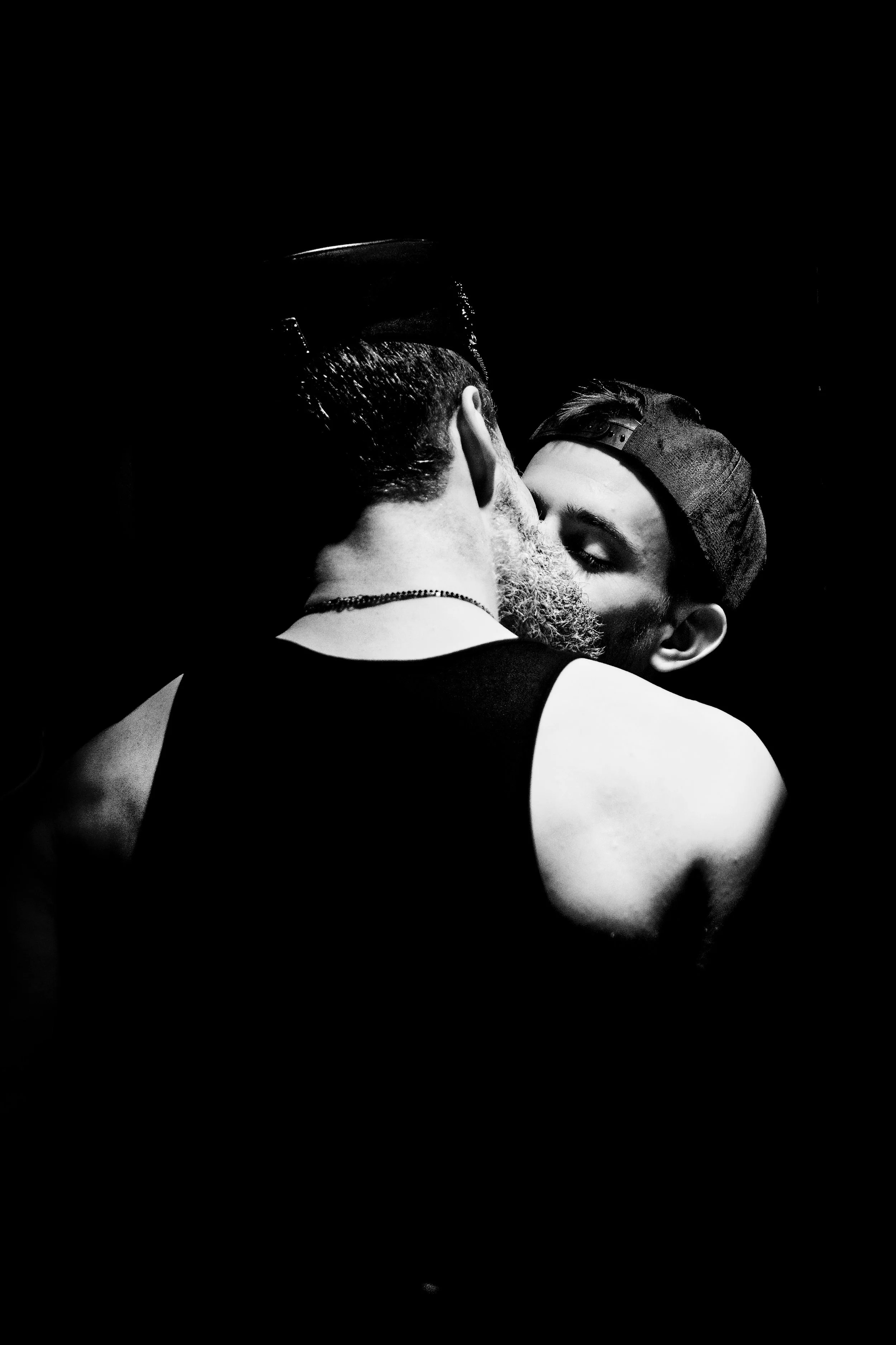 Two men, one with a beard and a cap, kissing in a black and white photograph against a dark background.