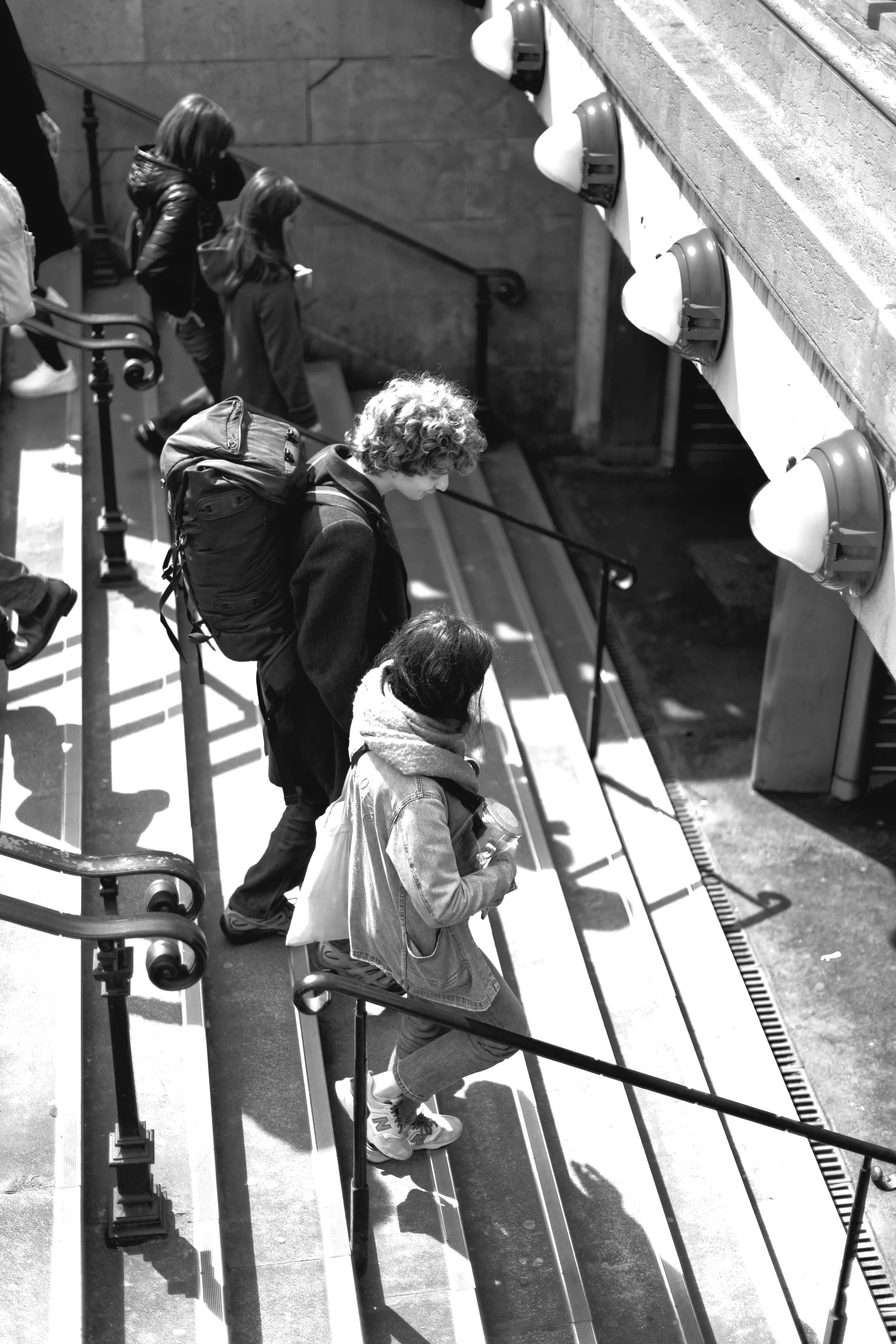 People waiting in line at a ticket booth with lamps overhead, sunlight casting shadows.