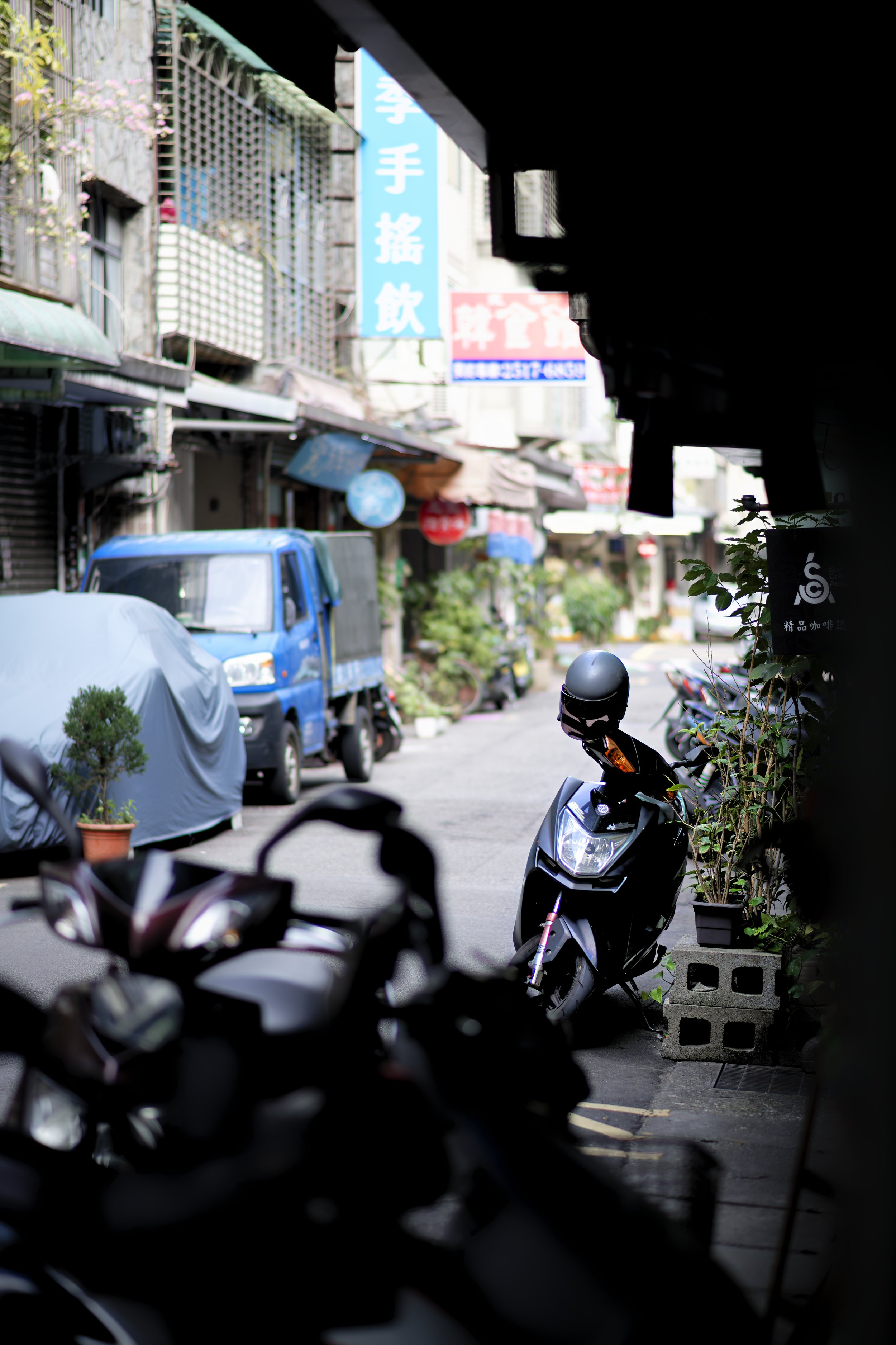 Quiet street scene with parked scooters and a blue truck, with buildings, plants, and signs in an Asian city