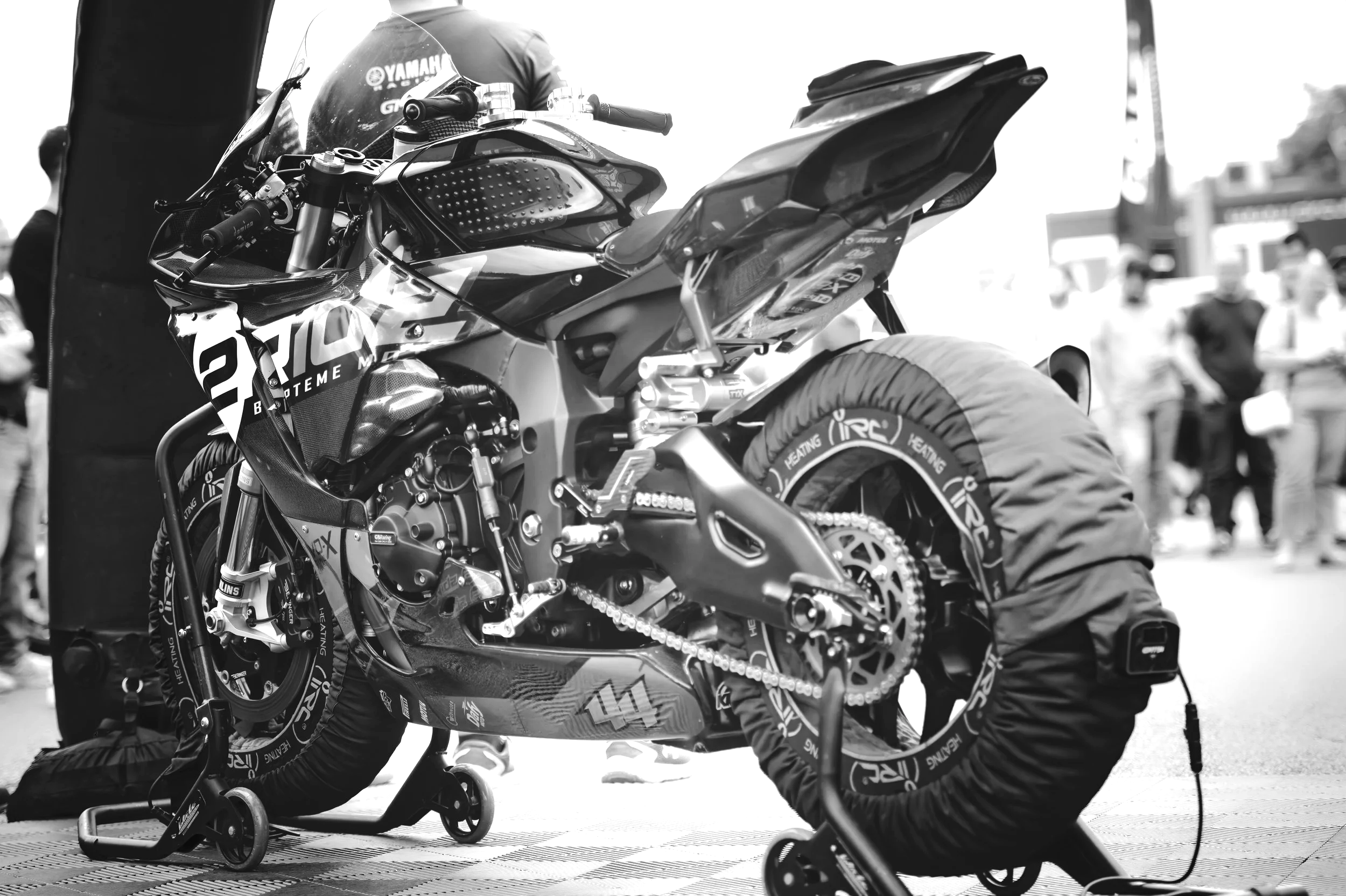 Black and white photo of a racing motorcycle on a paddock stand, with tires covered for protection, in a busy outdoor setting with several people in the background.