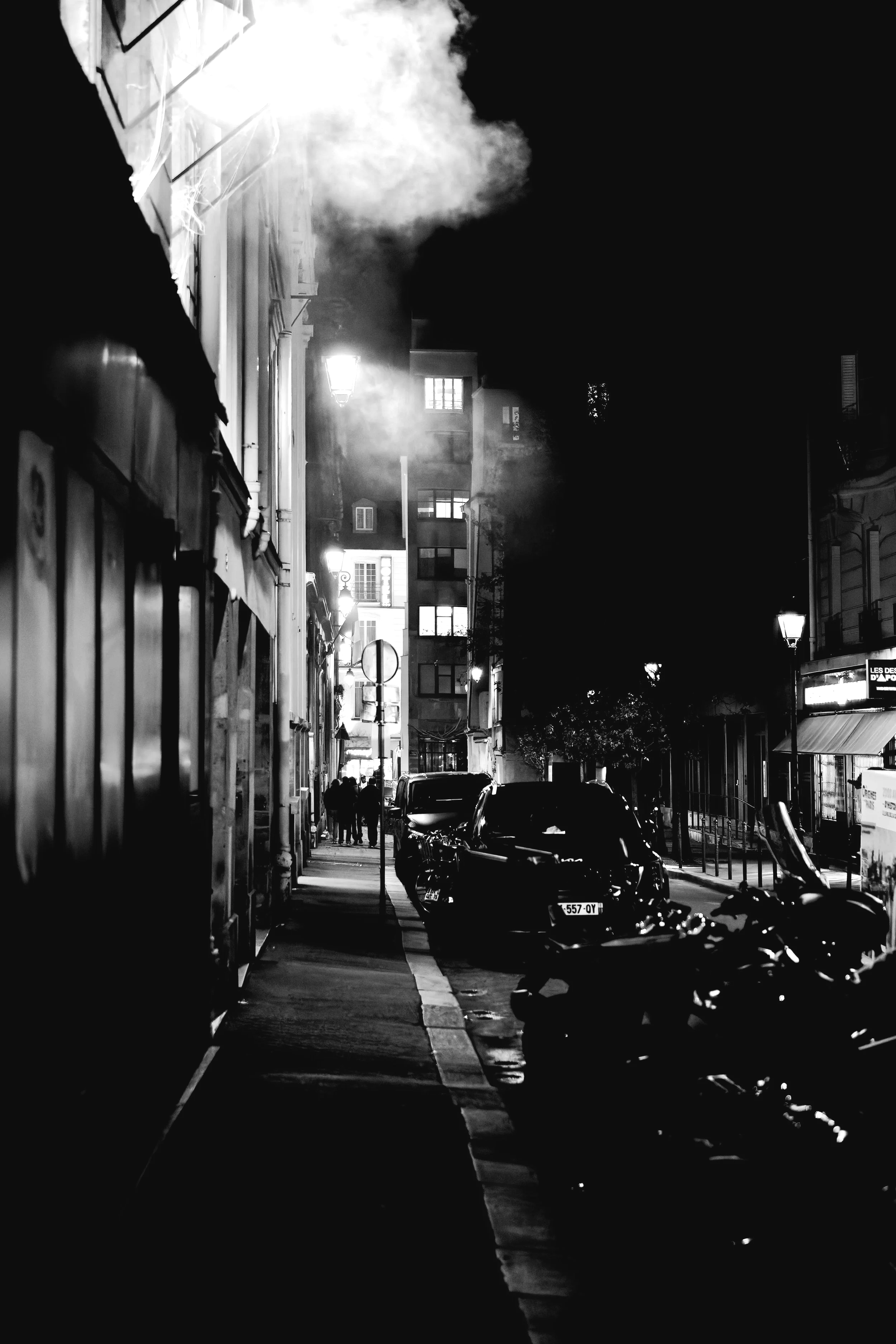 Nighttime city street scene in black and white with parked cars, motorcycles, illuminated street lamps, and building windows emitting light.