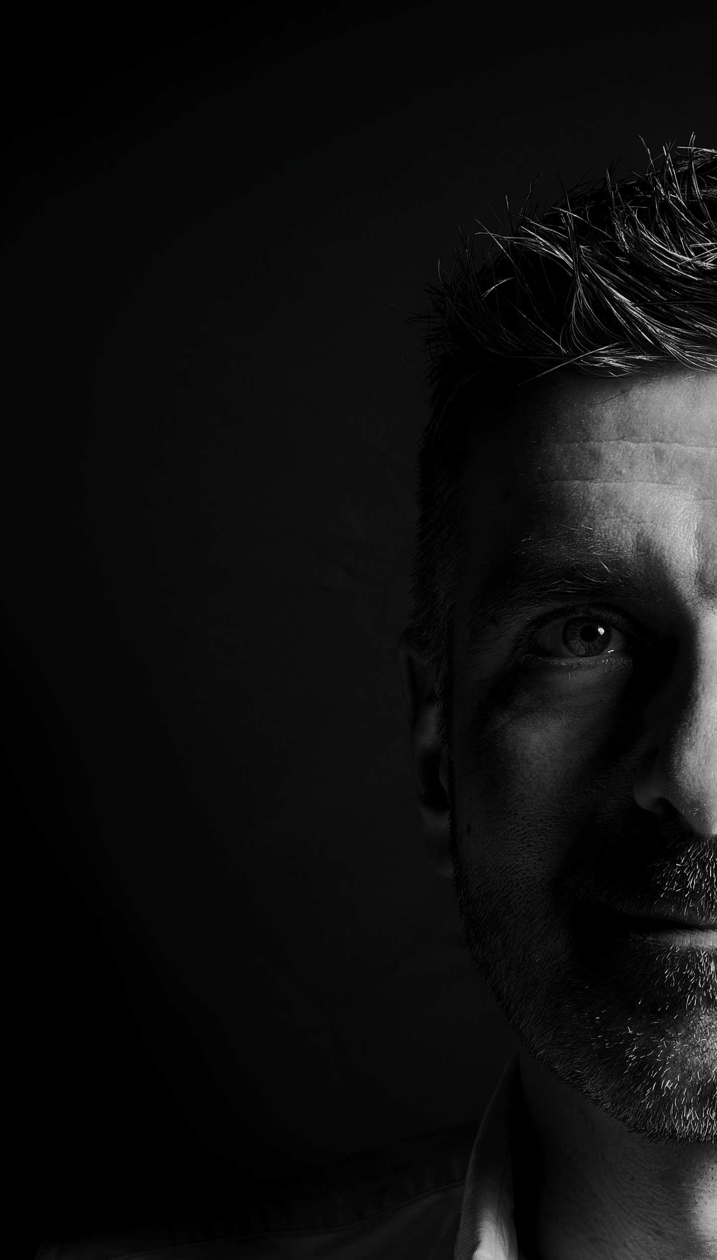 Black and white close-up photo of a man's face, half lit, with a beard and textured hair, looking directly at the camera.