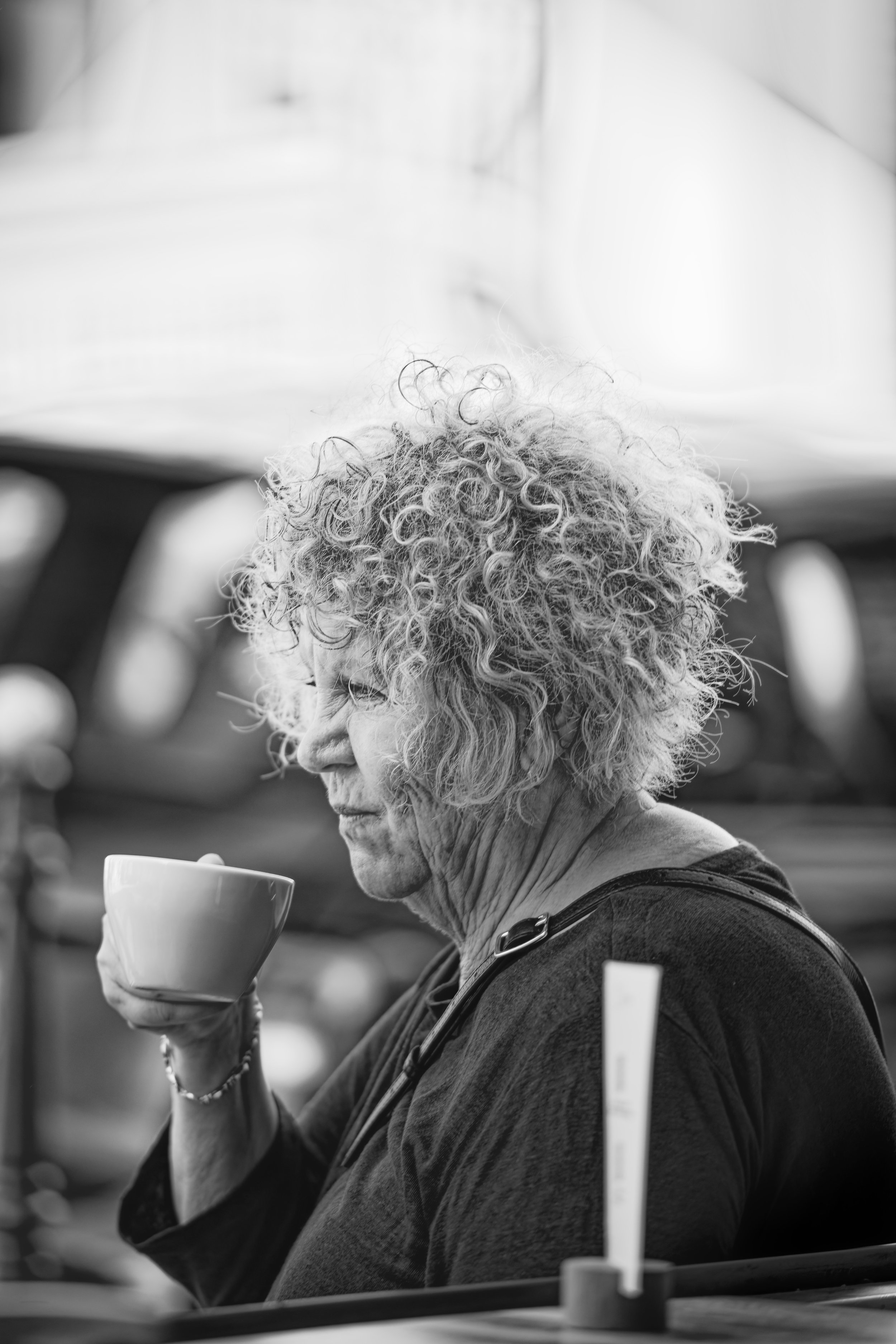 An elderly woman with curly hair drinking from a mug outdoors.