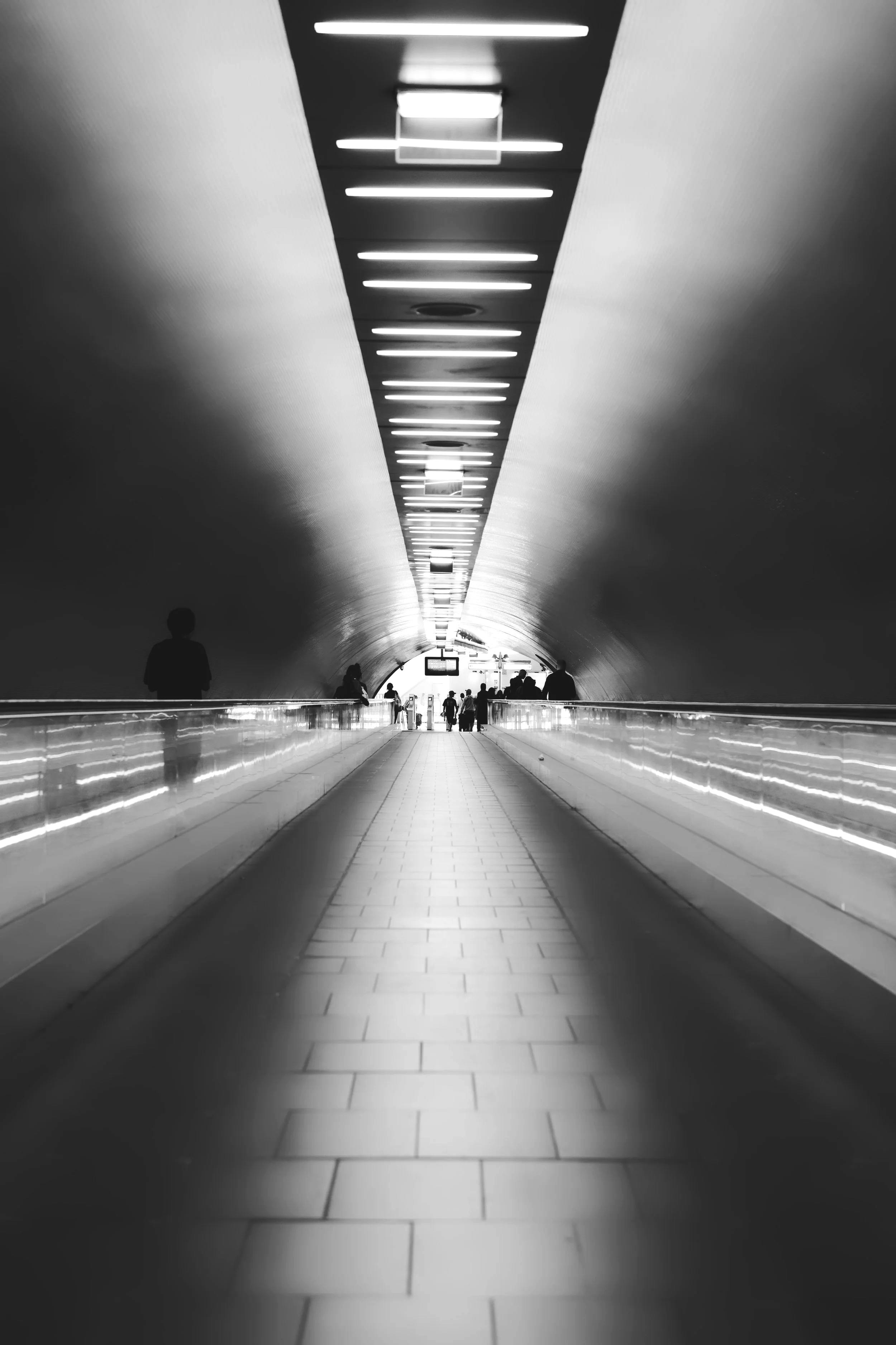 A black-and-white photo of an underground walkway with a ceiling lined with evenly spaced fluorescent lights, leading to an exit with silhouettes of people walking.