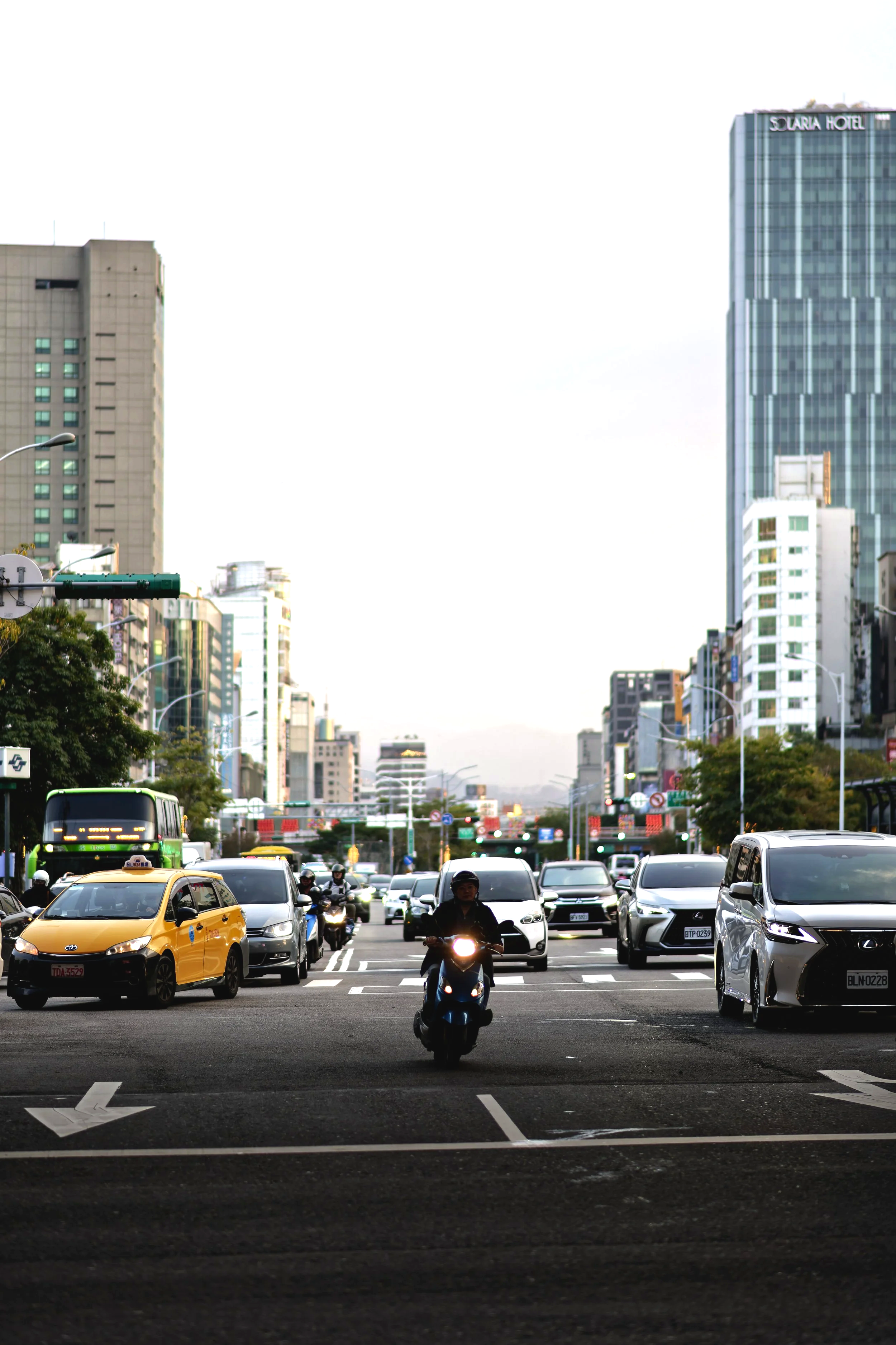 City street with cars, motorcycles, and tall buildings in the background, with a person riding a scooter in the center.
