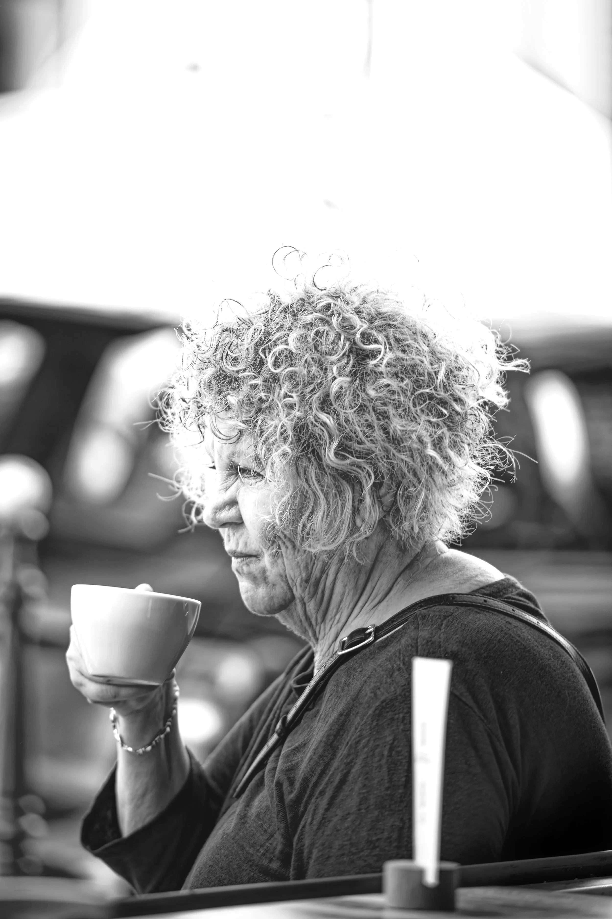 An elderly woman with curly hair drinking from a mug outdoors.