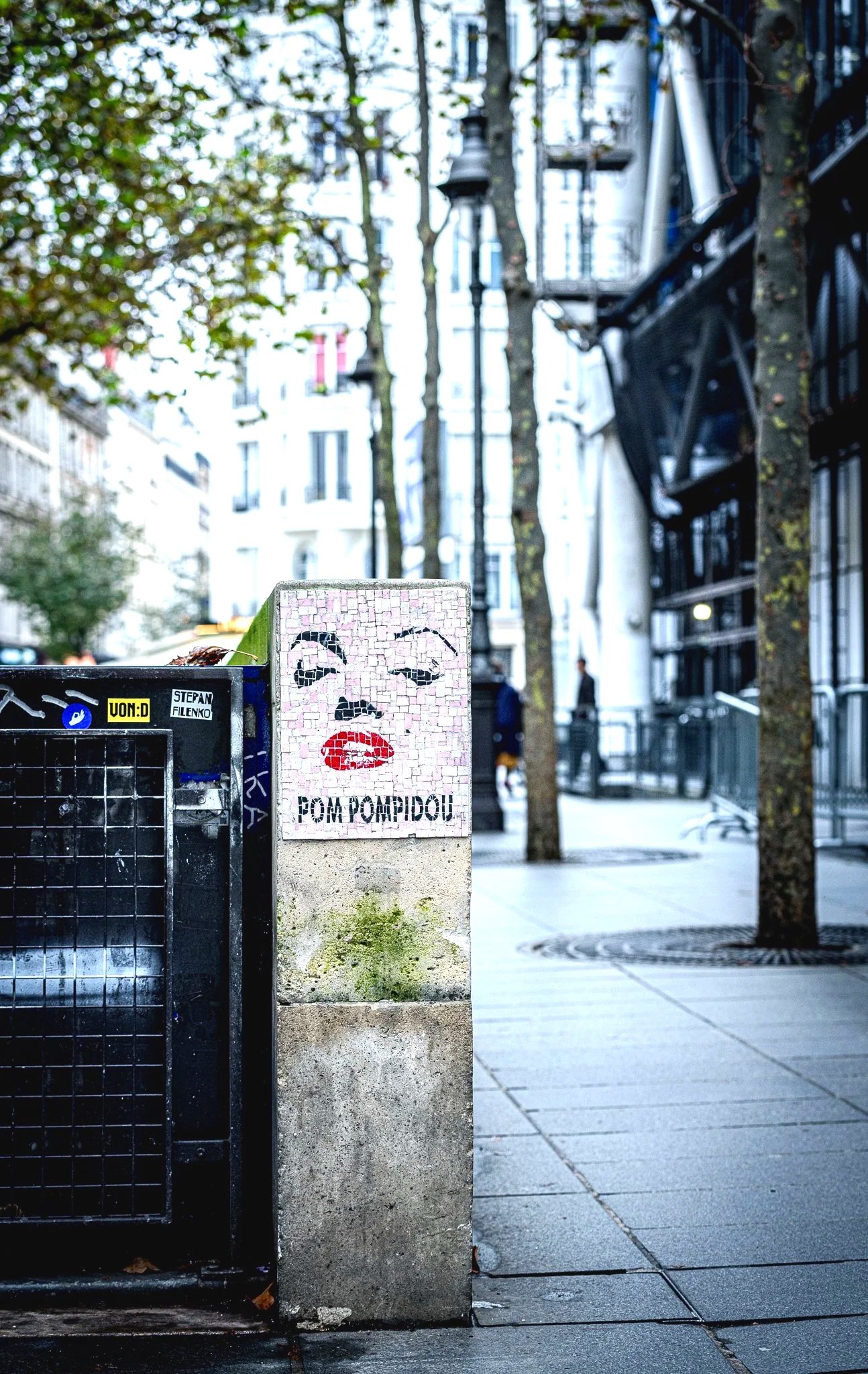 Street art mural of a woman's face with pink lips and black eyes, with The words POM POM PIDOU underneath. The mural is on a concrete pillar on a city sidewalk, with trees and buildings in the background.