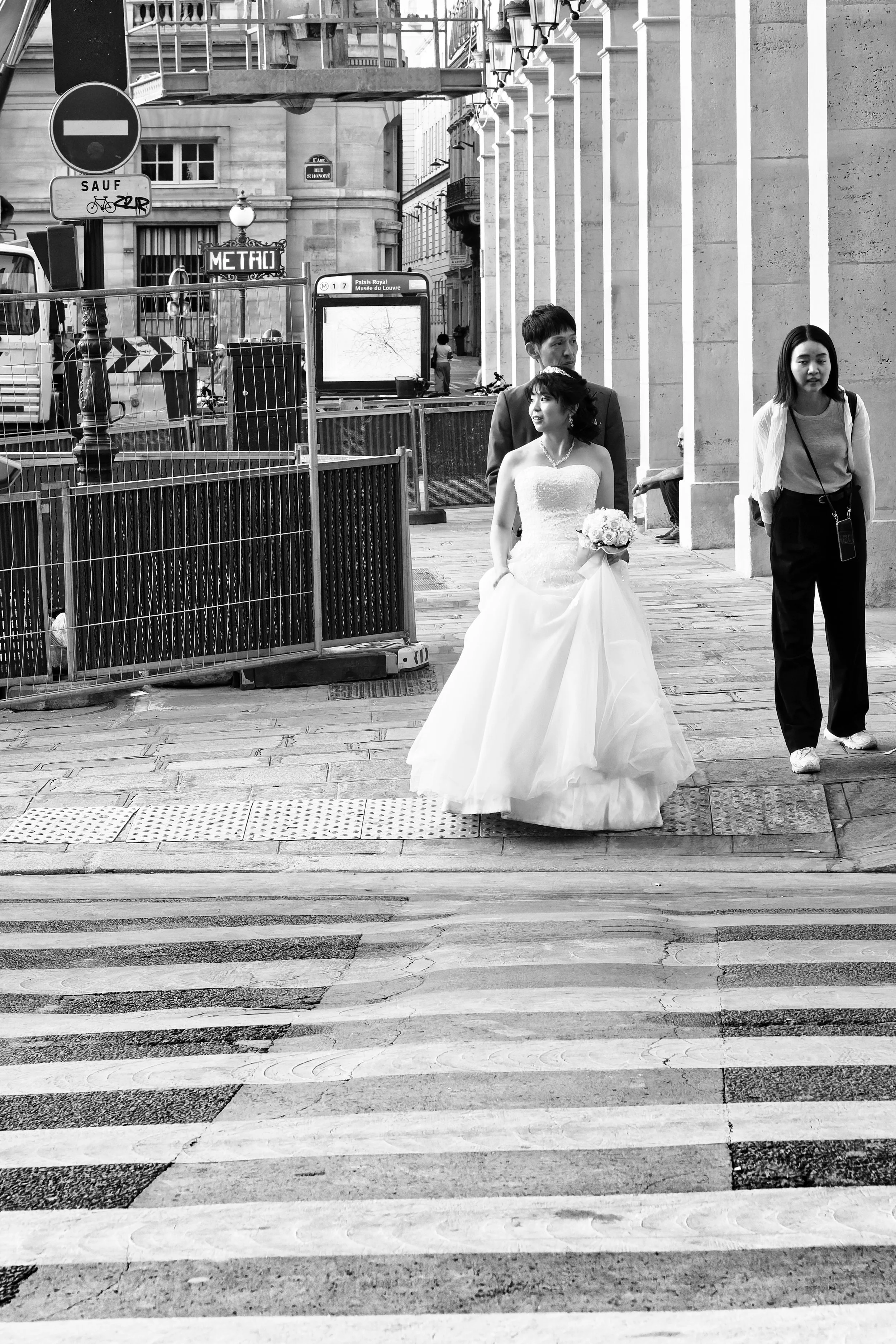 A bride in a wedding gown holding a bouquet walking across a crosswalk in an urban setting, accompanied by a man in a suit, with a woman nearby and city buildings in the background.