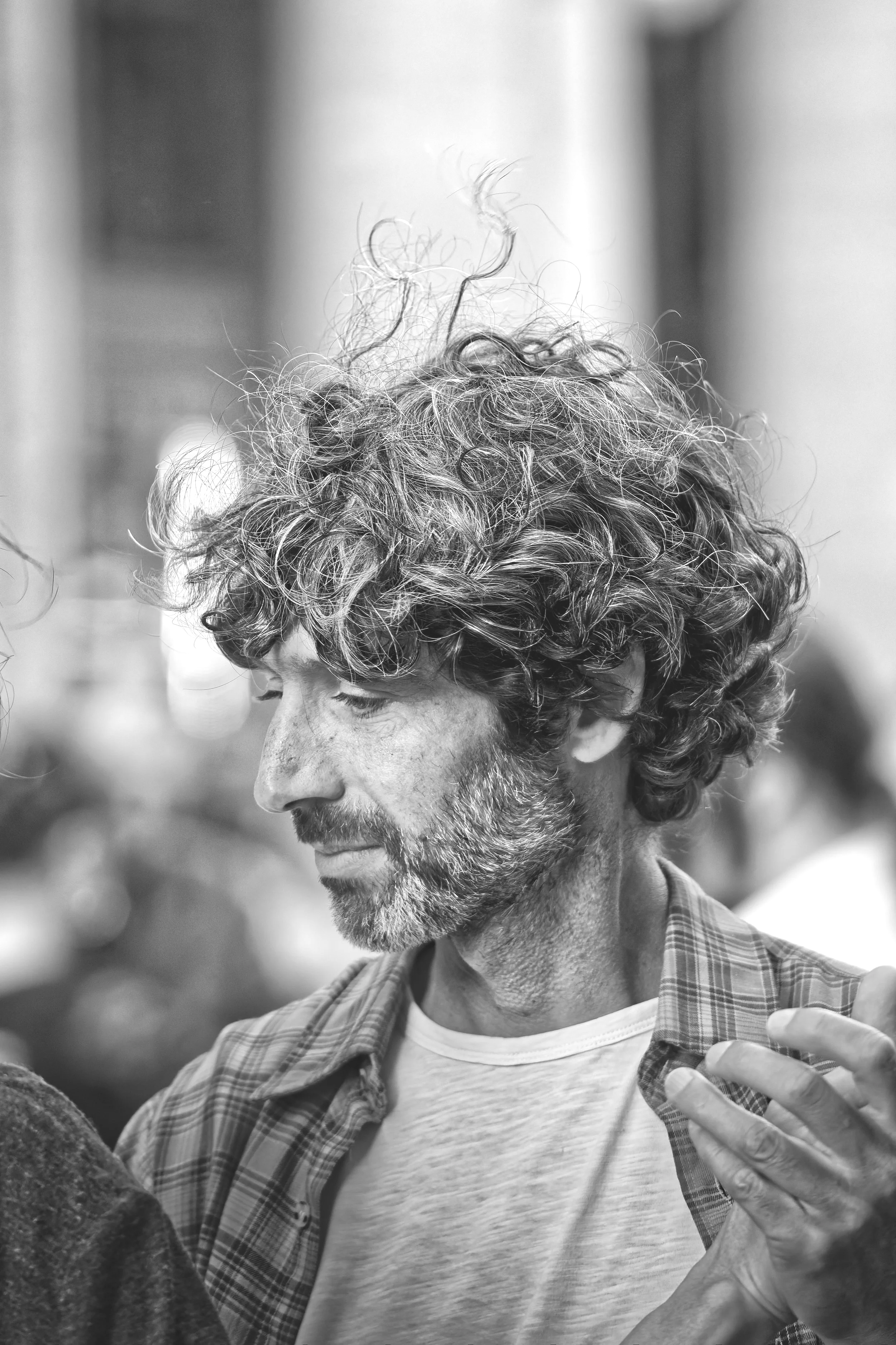 Black and white photo of a man with curly hair and a beard, wearing a plaid shirt and a white t-shirt, looking down with a calm expression.
