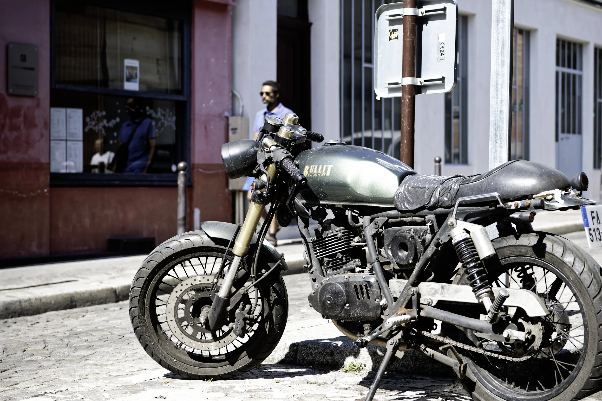 A vintage motorcycle with a green fuel tank and black seat parked on a cobblestone street, with a man in sunglasses walking in the background near buildings.