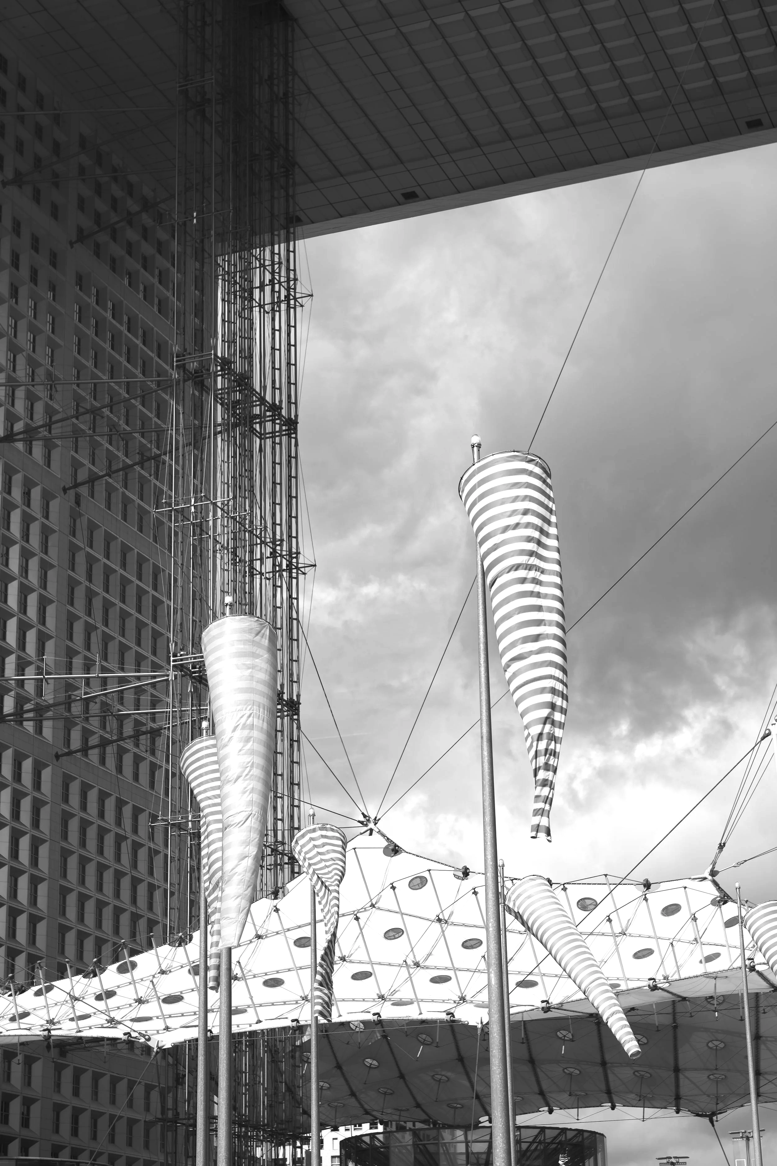 Black and white photo of striped kites flying near a large modern building and a structural canopy with lights.