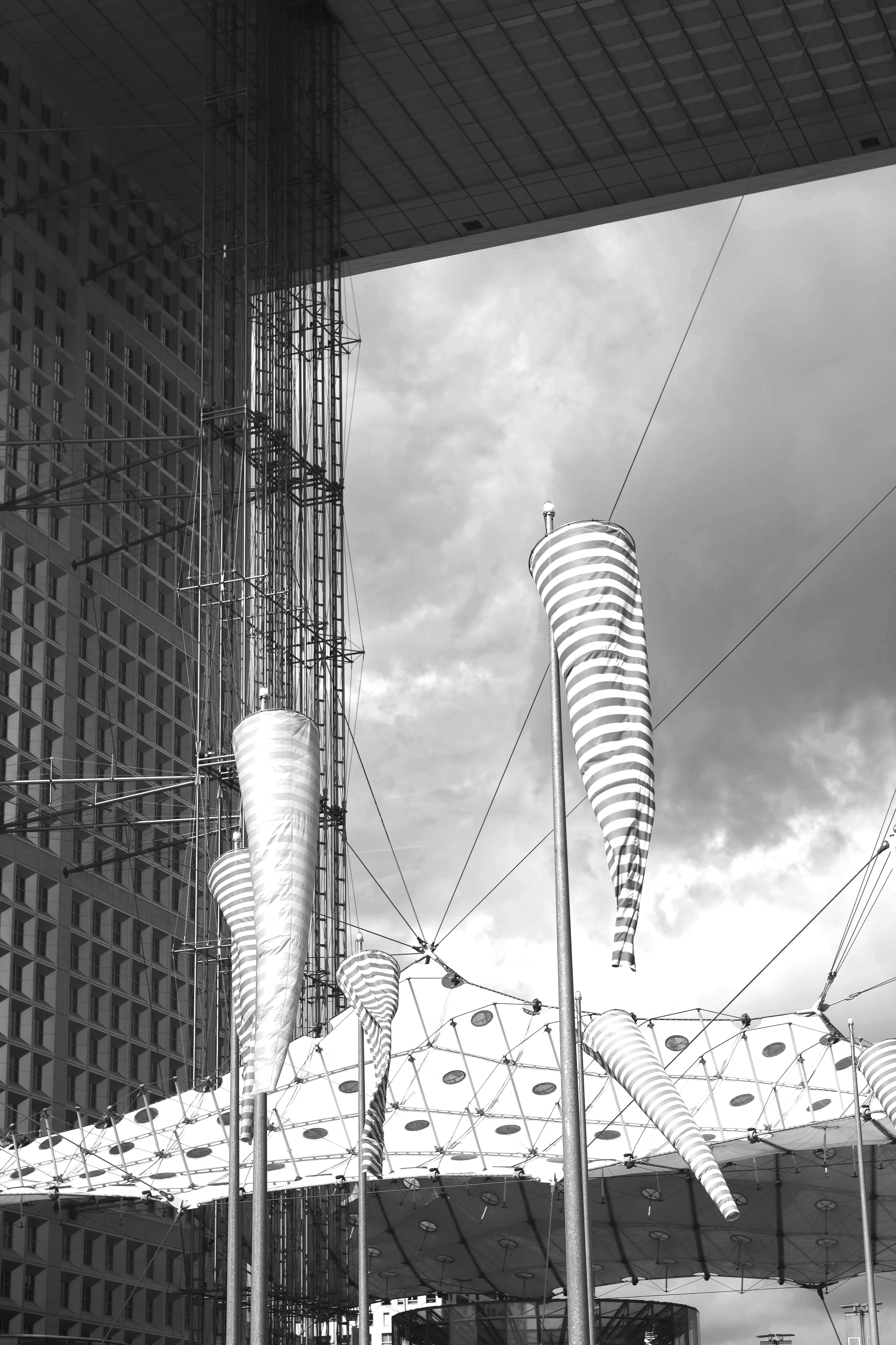 Black and white photo of striped kites flying near a large modern building and a structural canopy with lights.