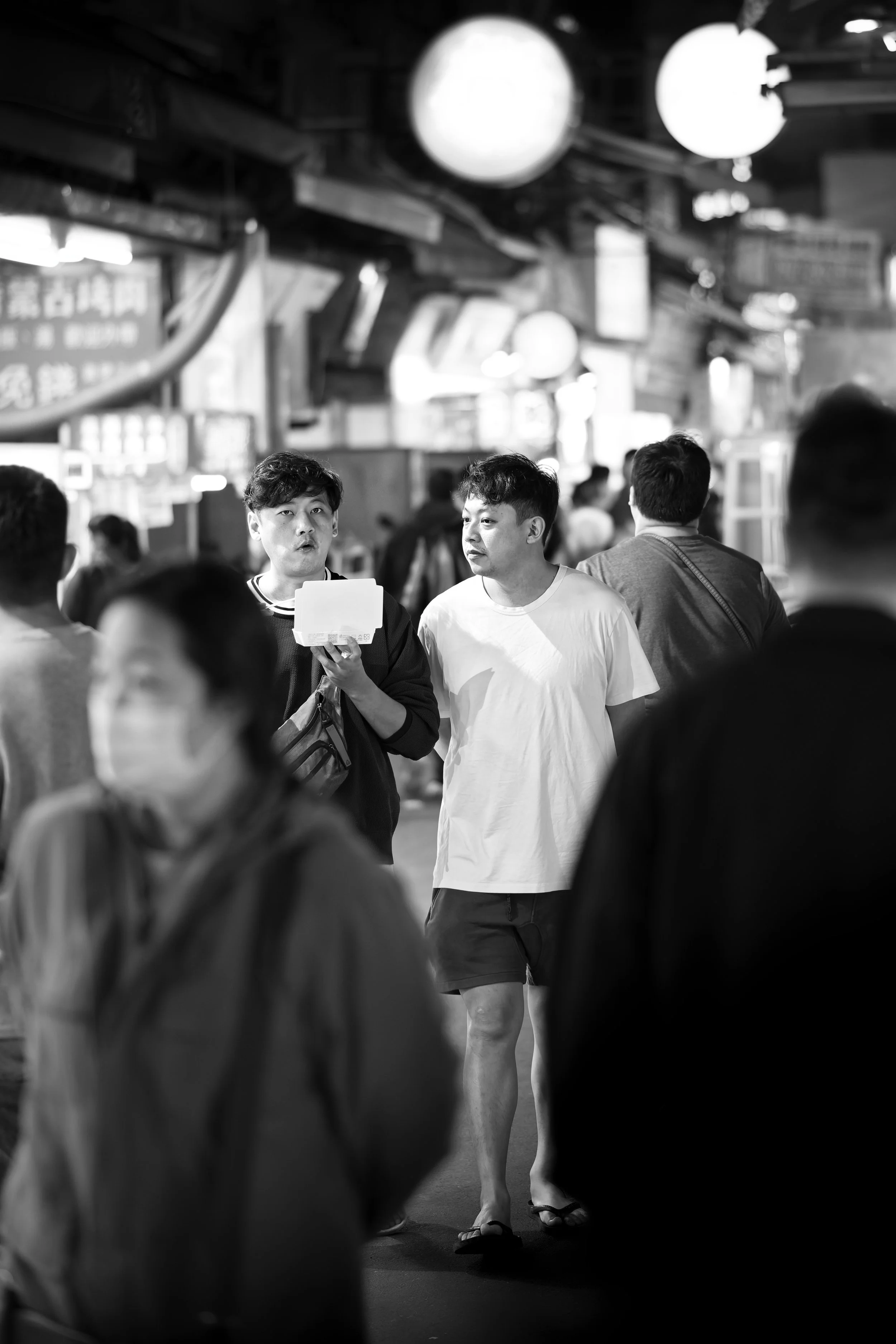 A black and white photo of a busy street scene with people walking, some wearing face masks, and two men in the center, one holding a box and the other looking at it, under illuminated street lights.
